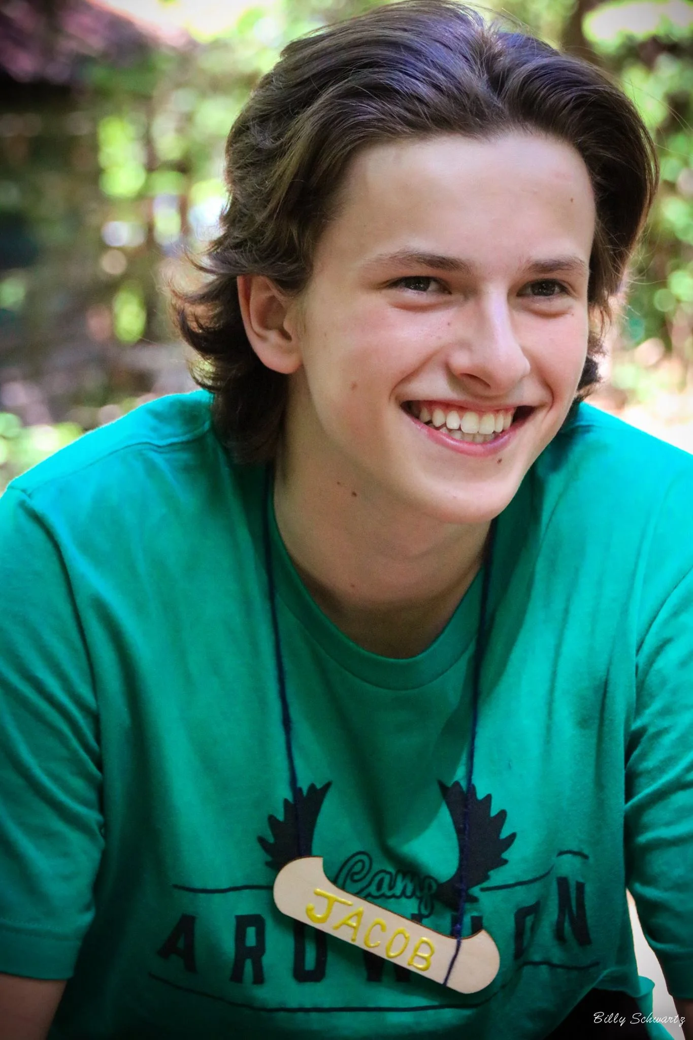 A young person with brown hair smiling, wearing a teal T-shirt with text and a name tag that reads 'JACOB' hanging around their neck, outdoors with a blurred green background.