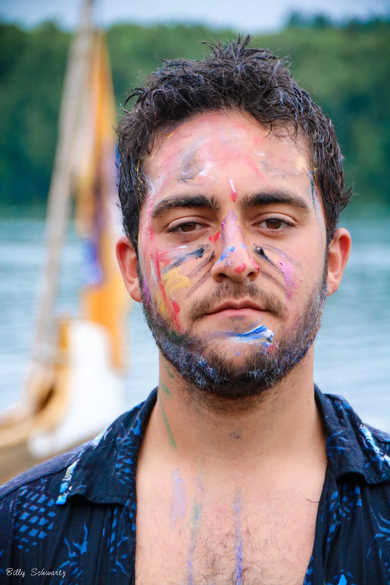 A young man with facial hair and short curly hair, with colorful paint smeared across his face and neck, standing outdoors near a body of water with trees in the background.