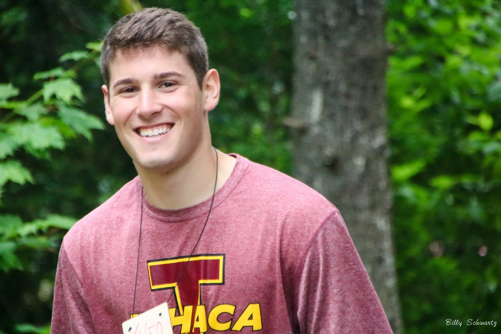 Smiling young man with short brown hair wearing a maroon T-shirt with a large yellow and red 'T' on the front, standing outdoors in a wooded area with green leaves and trees in the background.