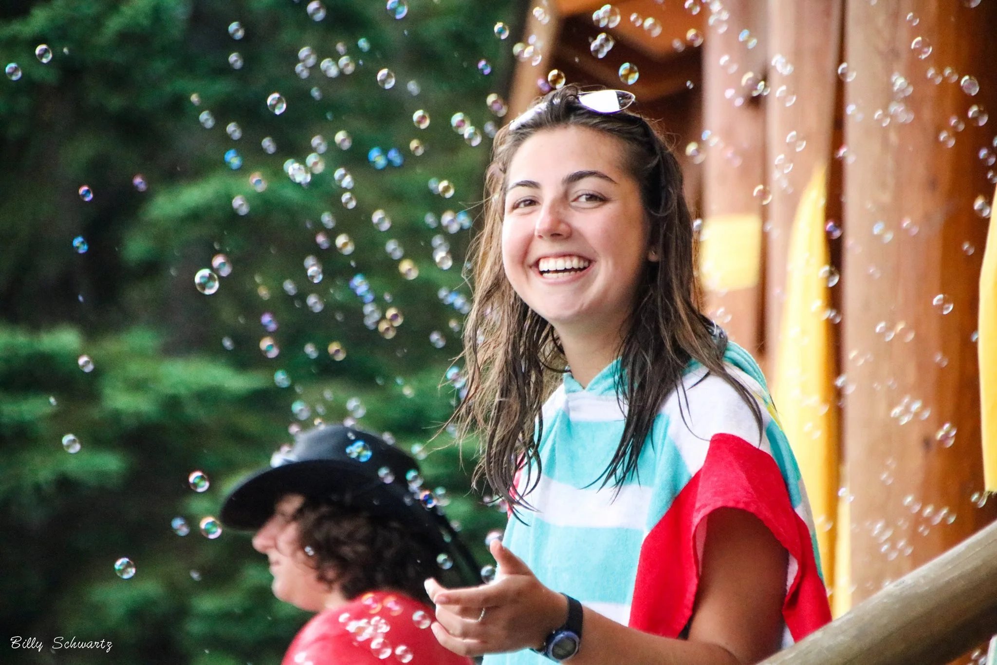 Young woman smiling with wet hair and sunglasses on her head, surrounded by soap bubbles outdoors with a wooden background and someone in a black hat nearby.