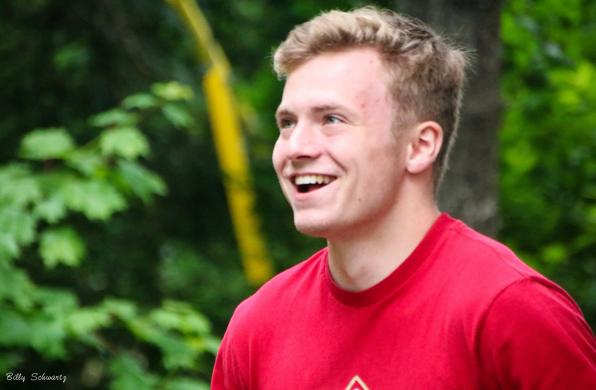 A young man with short, curly blonde hair and fair skin, smiling enthusiastically, wearing a red shirt, standing outdoors in front of lush green trees and leaves.