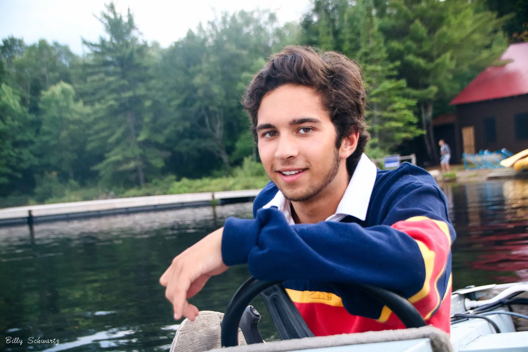 A young man with dark brown curly hair and a light beard, wearing a colorful jacket, smiling at the camera while sitting on a boat by the lake with green trees in the background.