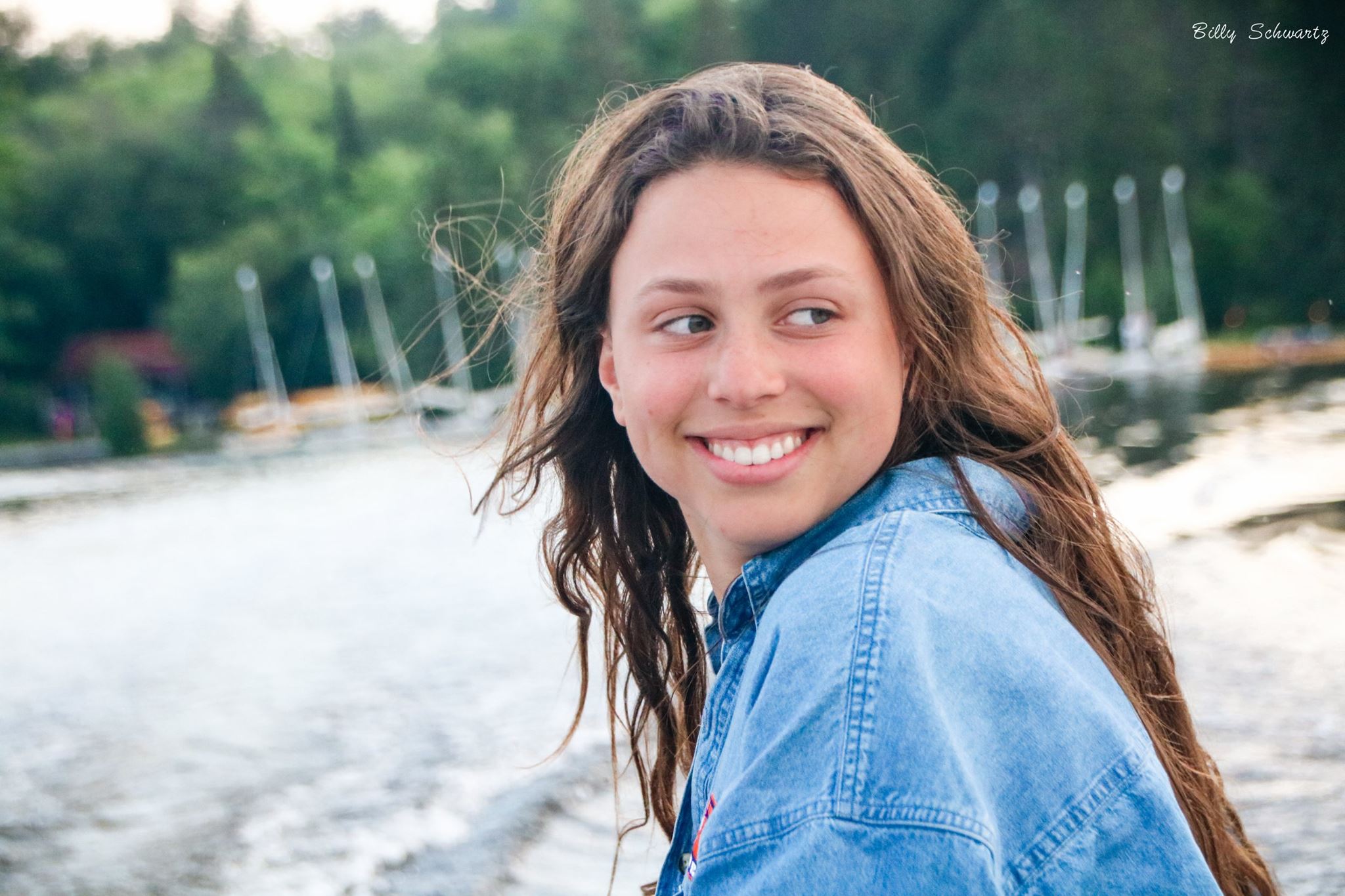 A young woman with long brown hair smiling and looking to her left, sitting near a body of water with boats and trees in the background.