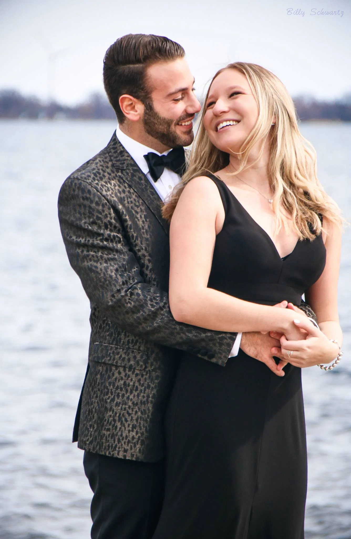 A man and woman embrace near a body of water, smiling and looking at each other. The man is wearing a tuxedo with a leopard print jacket and a bow tie. The woman is wearing a black sleeveless dress. Both appear happy and are close together.