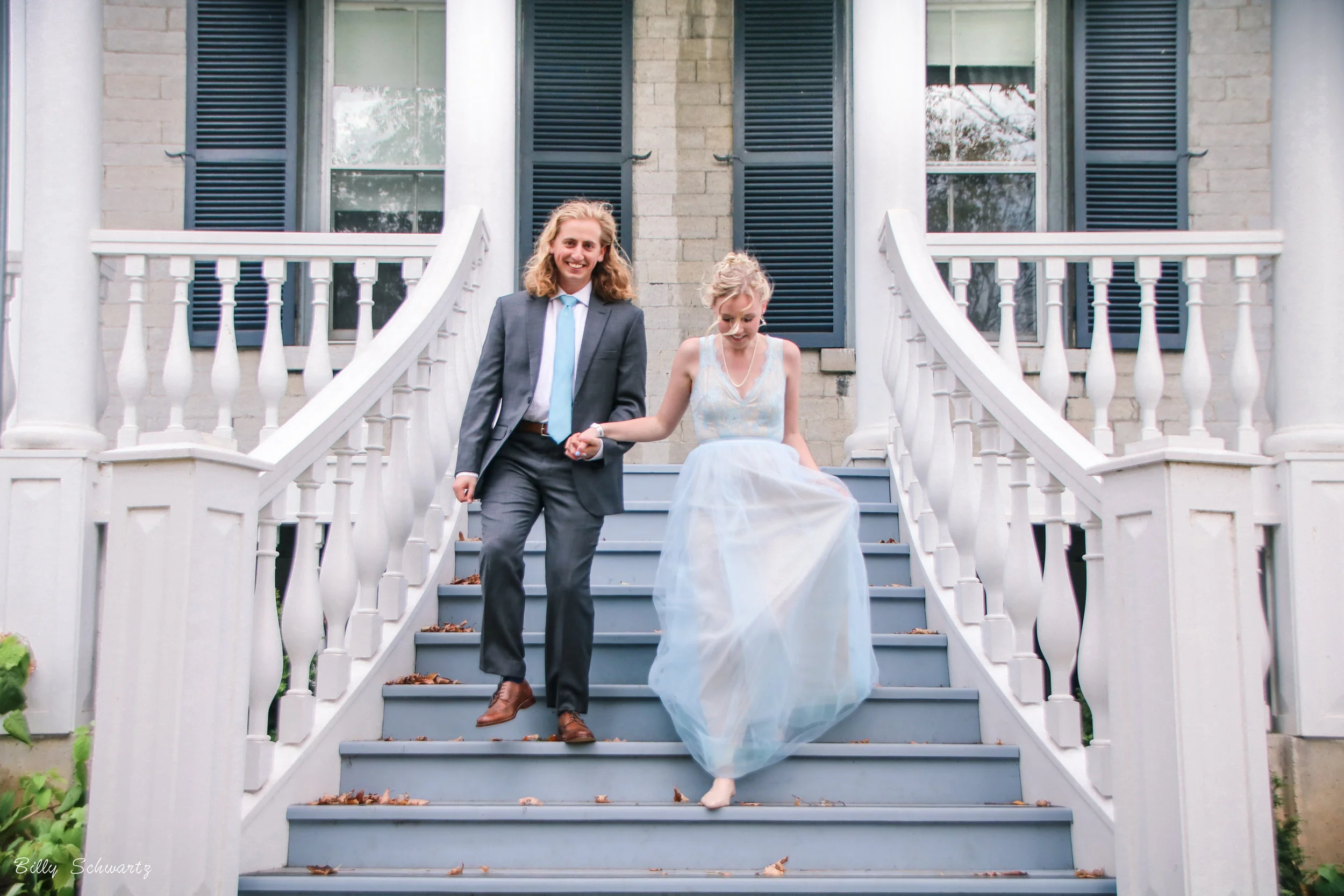 A man in a suit helping a woman in a white dress down the steps of a porch with blue railings and white columns.