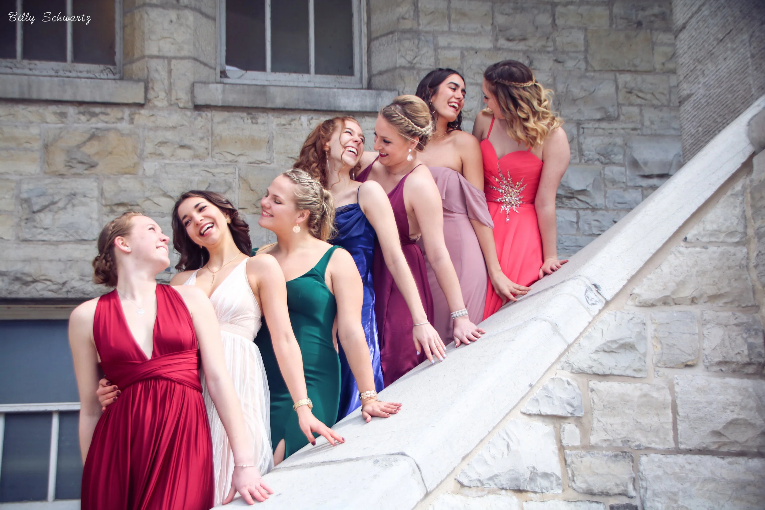 Group of nine young women in colorful evening gowns standing on a stone staircase, smiling and interacting with each other.