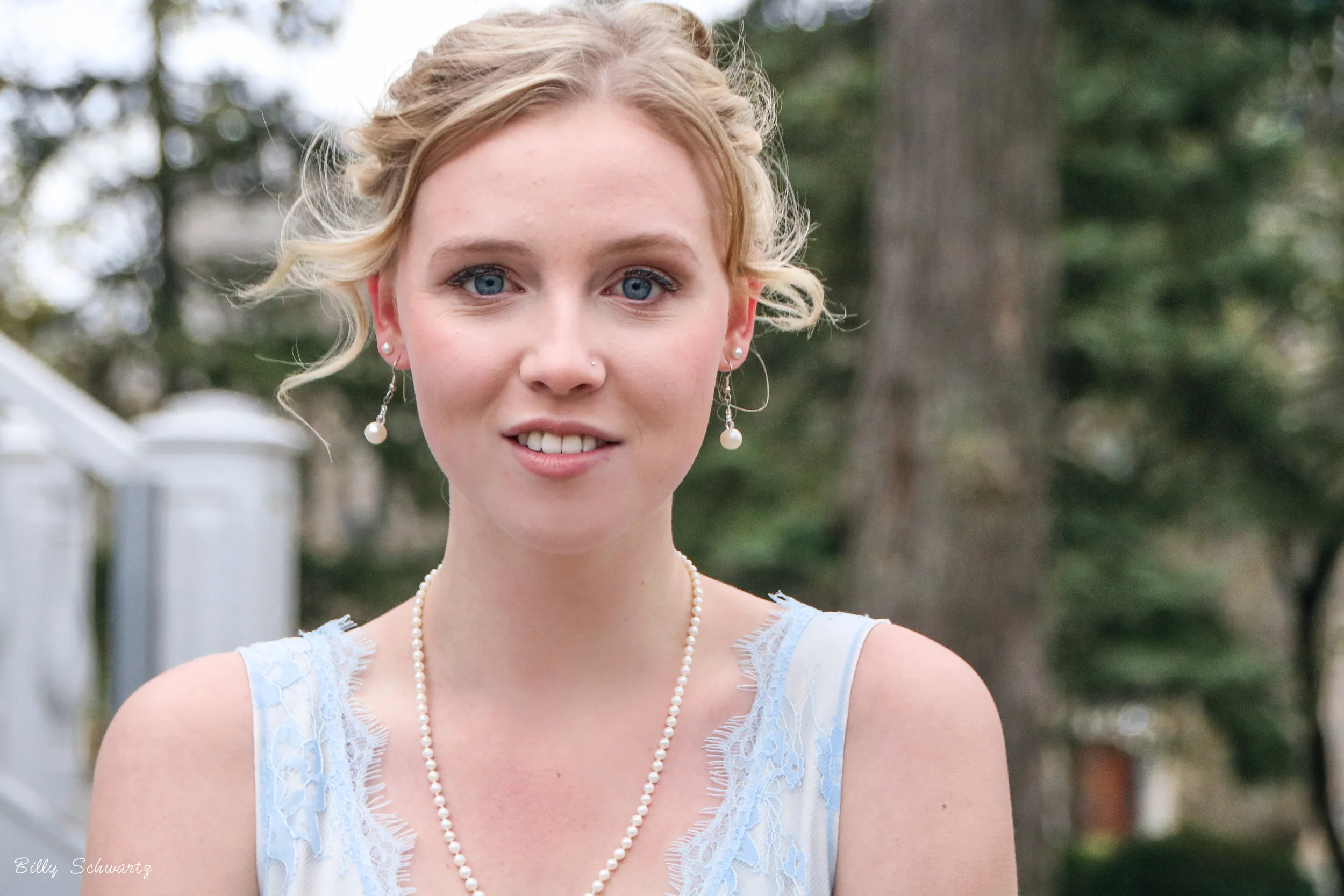 A young woman with blonde hair styled in curls, wearing pearl earrings, a pearl necklace, and a white dress with light blue lace, standing outdoors with trees in the background.