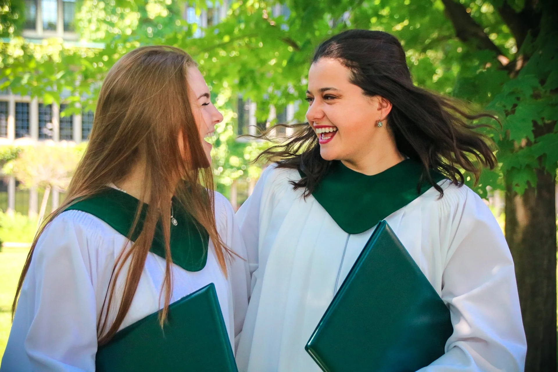 Two young women in graduation gowns smiling and laughing outdoors, holding green diplomas, with trees and a building in the background.