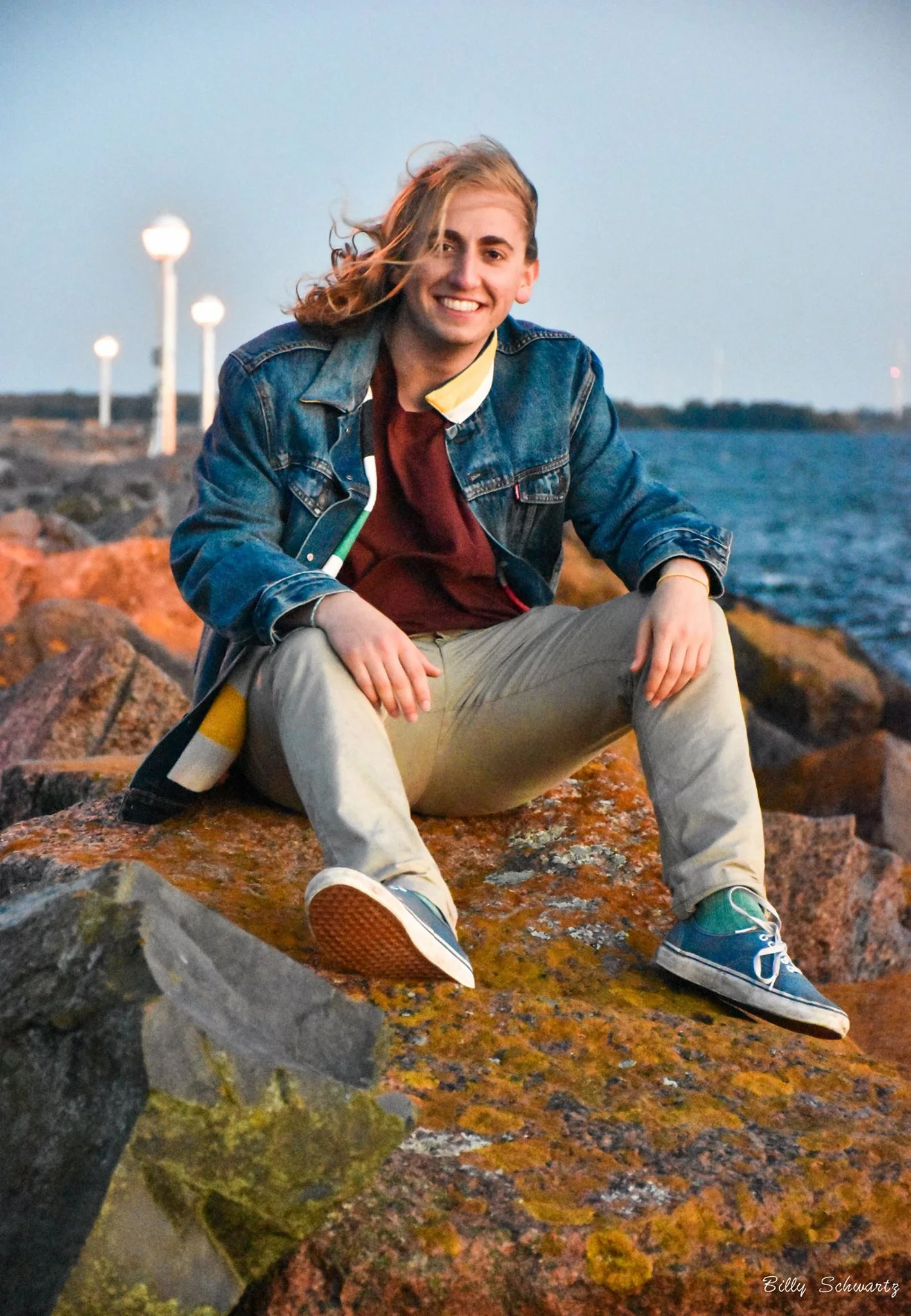 A young man with long, wavy blonde hair, sitting on a large orange-brown rock by the water with a smile on his face, wearing a denim jacket, red shirt, beige pants, and blue sneakers, during sunset.
