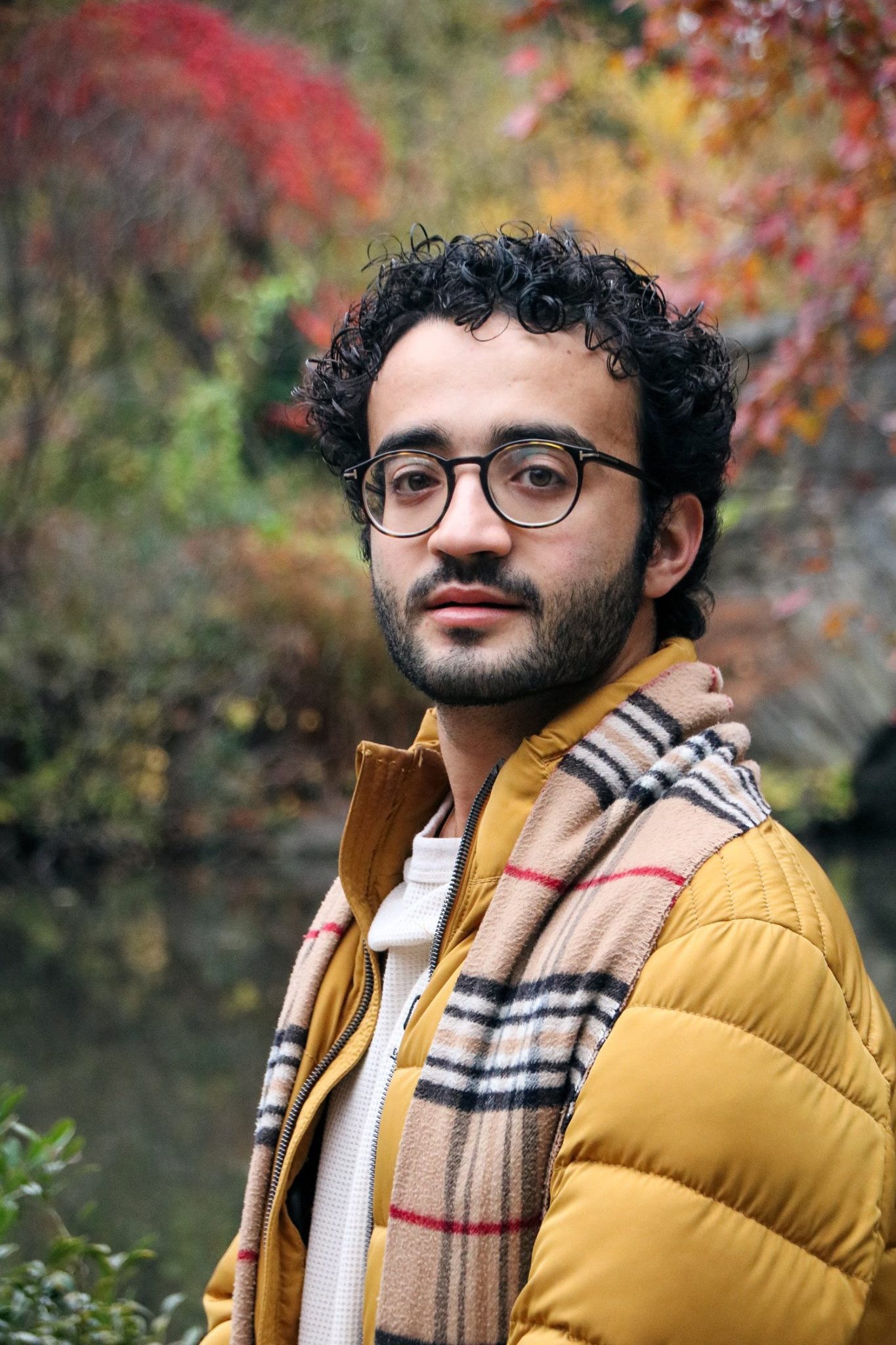 A man with curly dark hair and glasses stands outdoors during autumn near a body of water, wearing a mustard yellow quilted jacket and a beige plaid scarf.