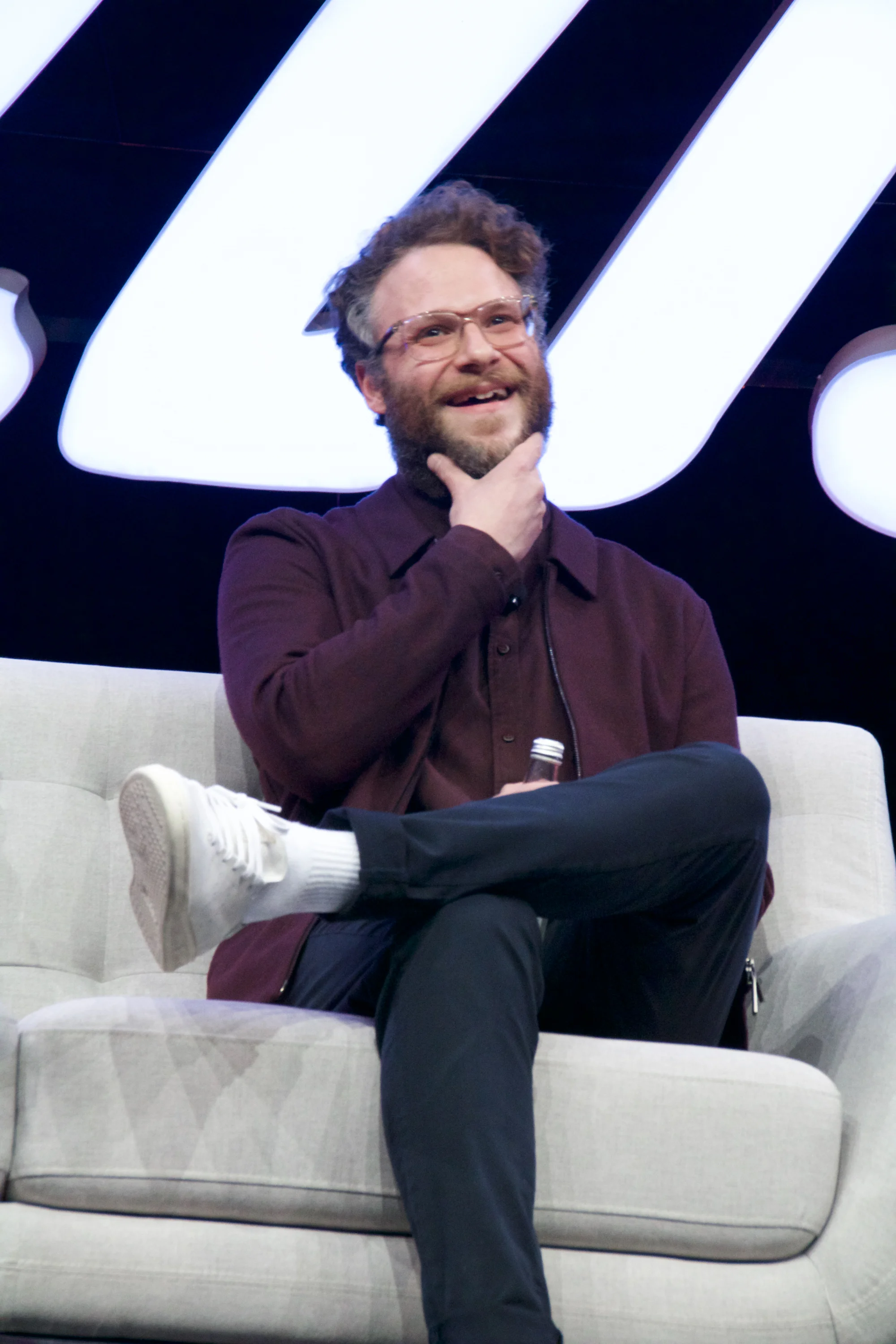 A man with glasses and a beard, wearing a maroon jacket and black pants, sitting on a beige couch with one leg crossed over the other, smiling and touching his chin, holding a small bottle in his hand, in front of a large illuminated sign.