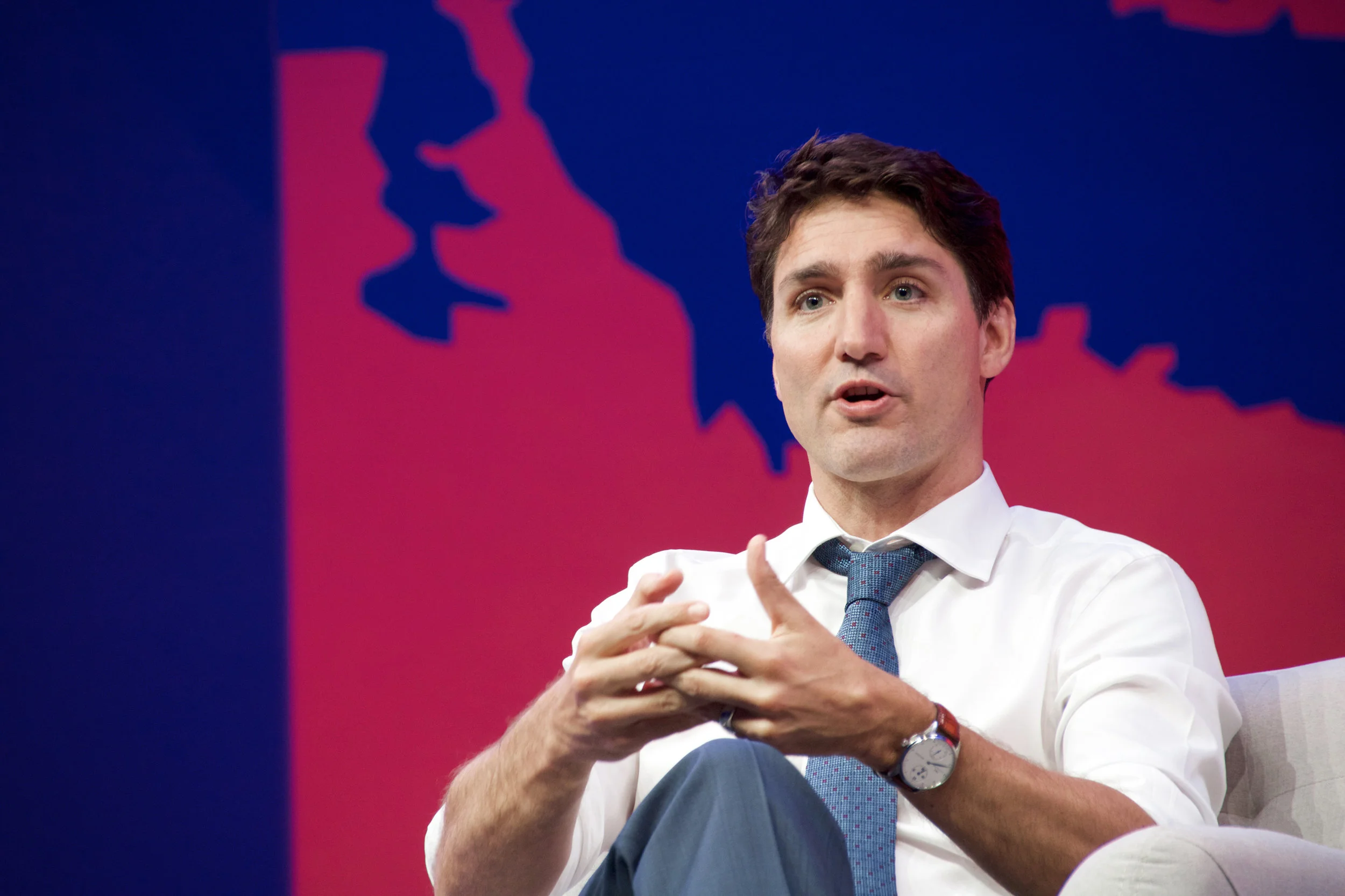 Man in white shirt and blue tie speaking, sitting on a chair, with a red and blue background featuring a silhouette of a map.