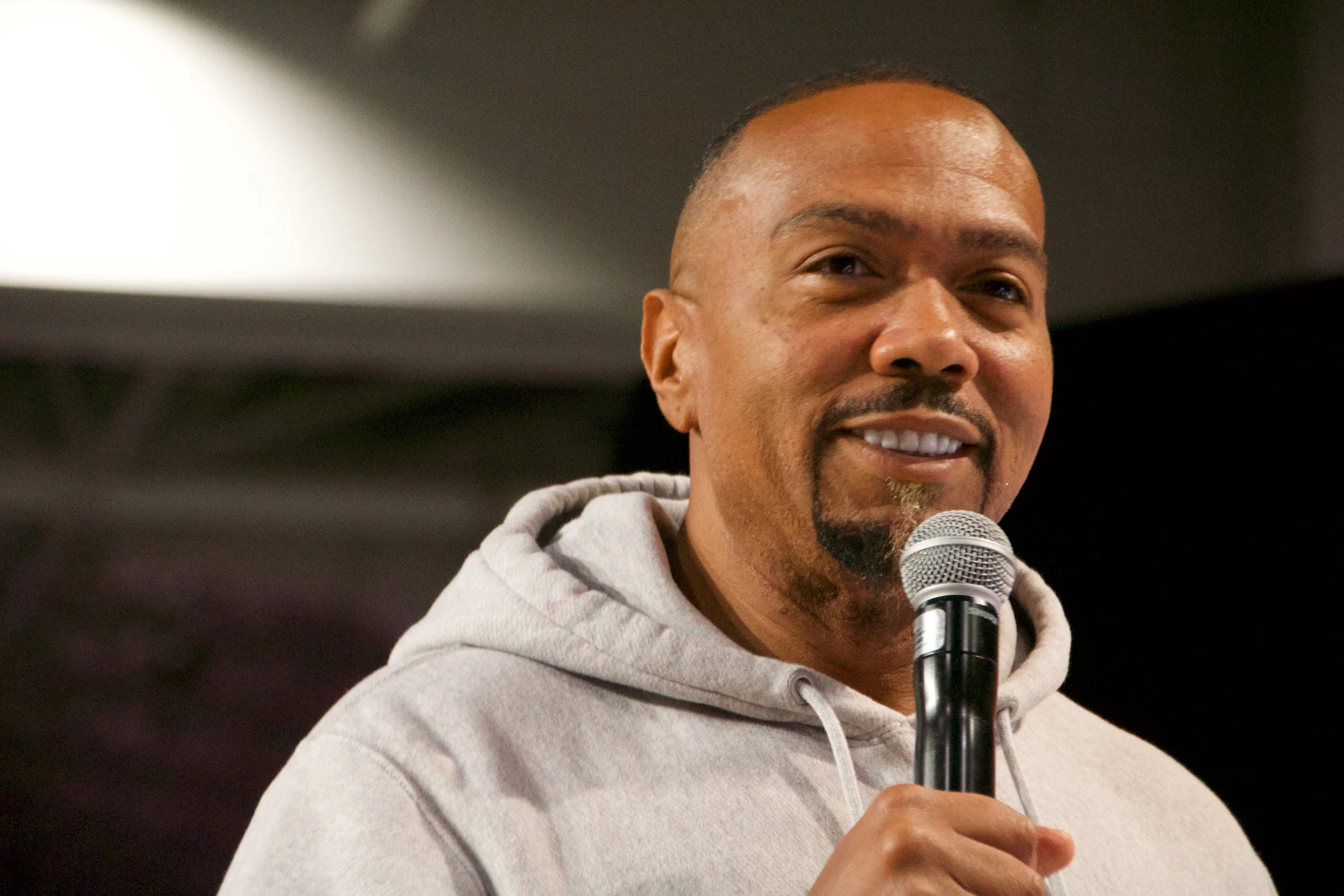 A man smiling and holding a microphone, wearing a light gray hoodie.