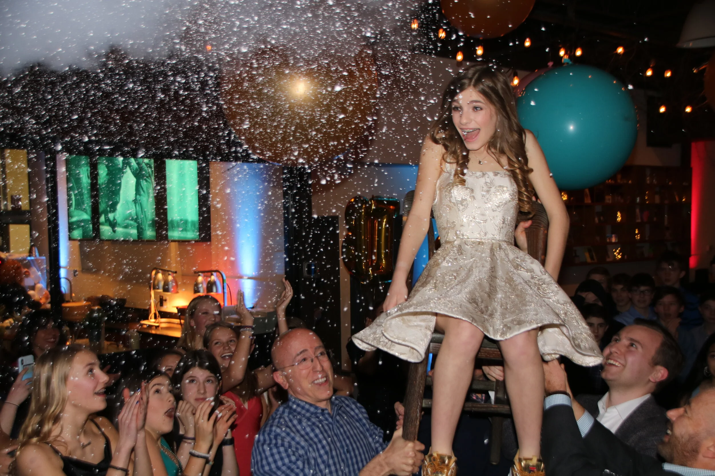 Girl in a gold dress on a chair during a celebration with people surrounding her, throwing confetti, in a decorated room with balloons and lights.