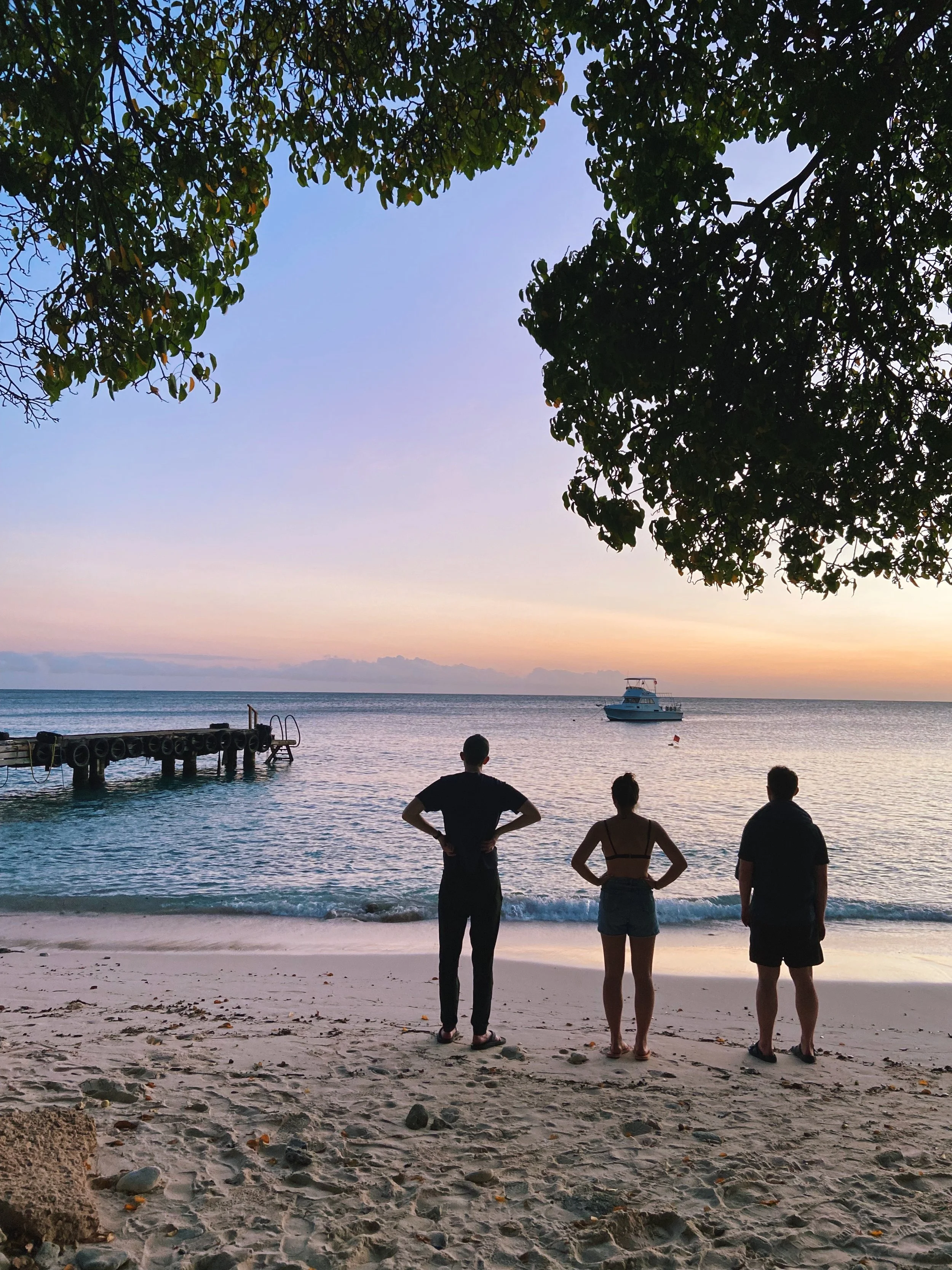 Three people standing on a sandy beach, gazing at the sunset over the ocean, with trees overhead and a boat in the water near a small dock.