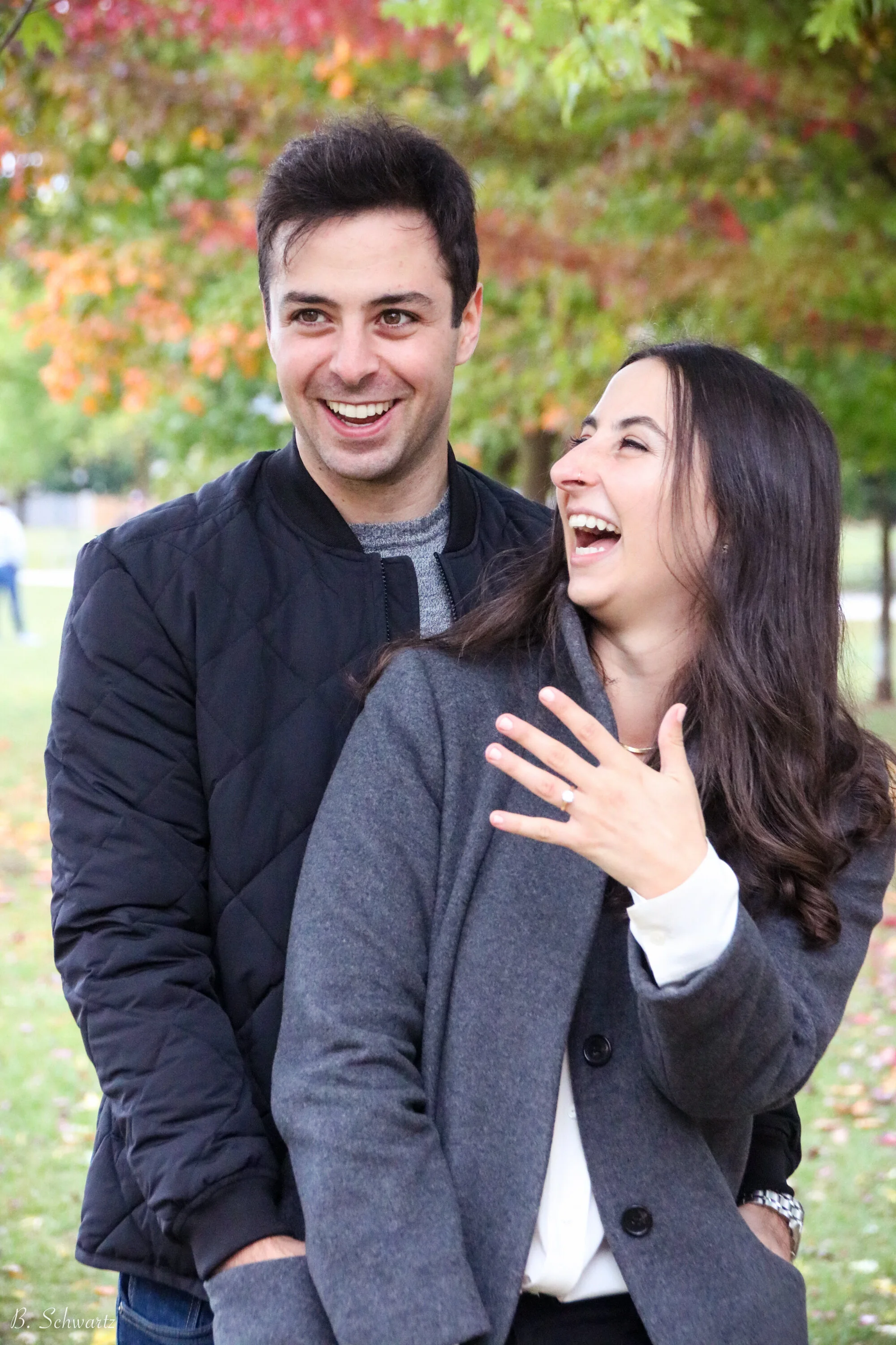 A man and woman laughing and smiling outdoors in a park with trees displaying fall foliage.