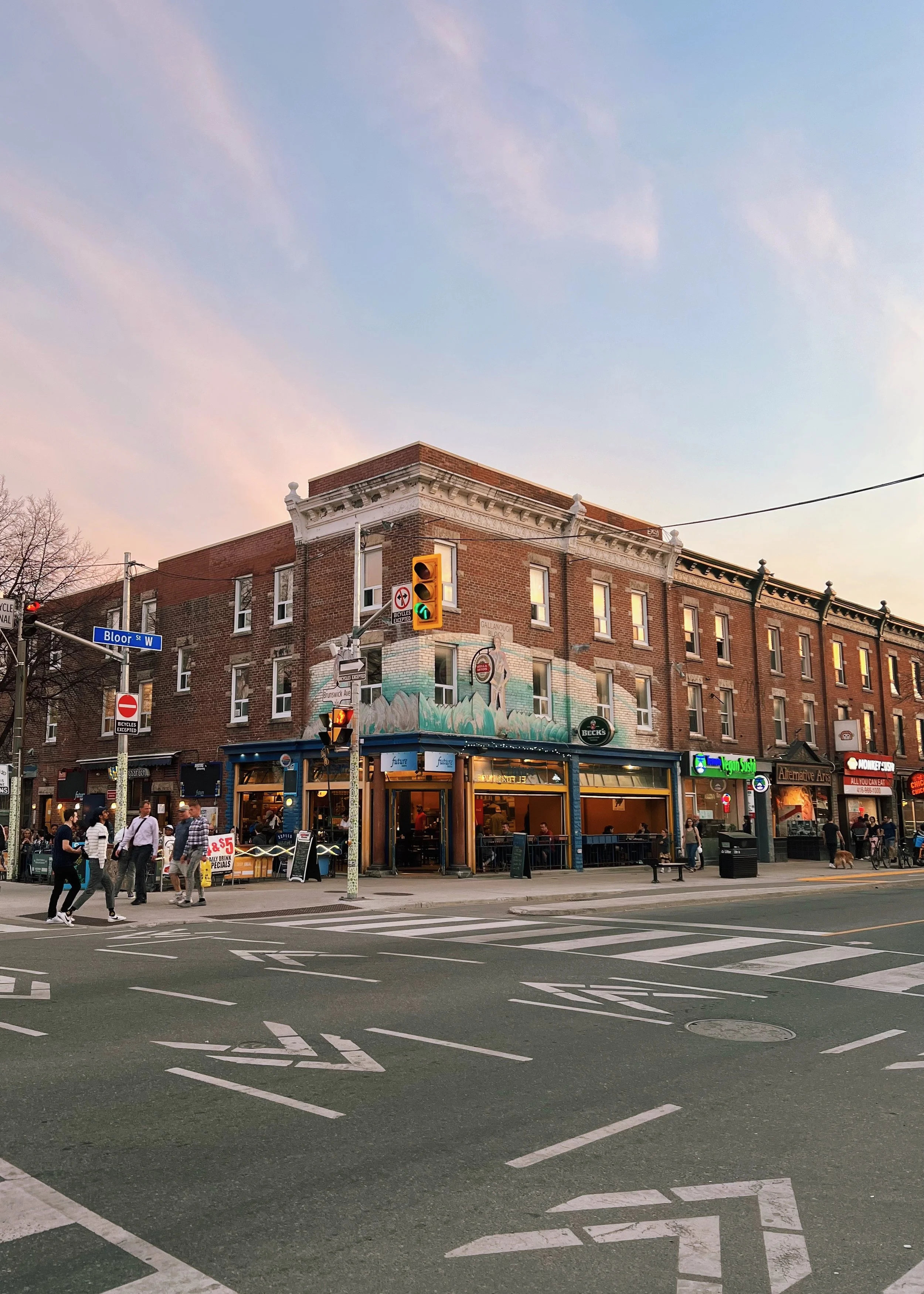 Street corner with a three-story brick building housing a restaurant, bars, or pub on the ground floor. Pedestrians crossing and walking along the sidewalk during sunset with traffic lights and street signs visible.