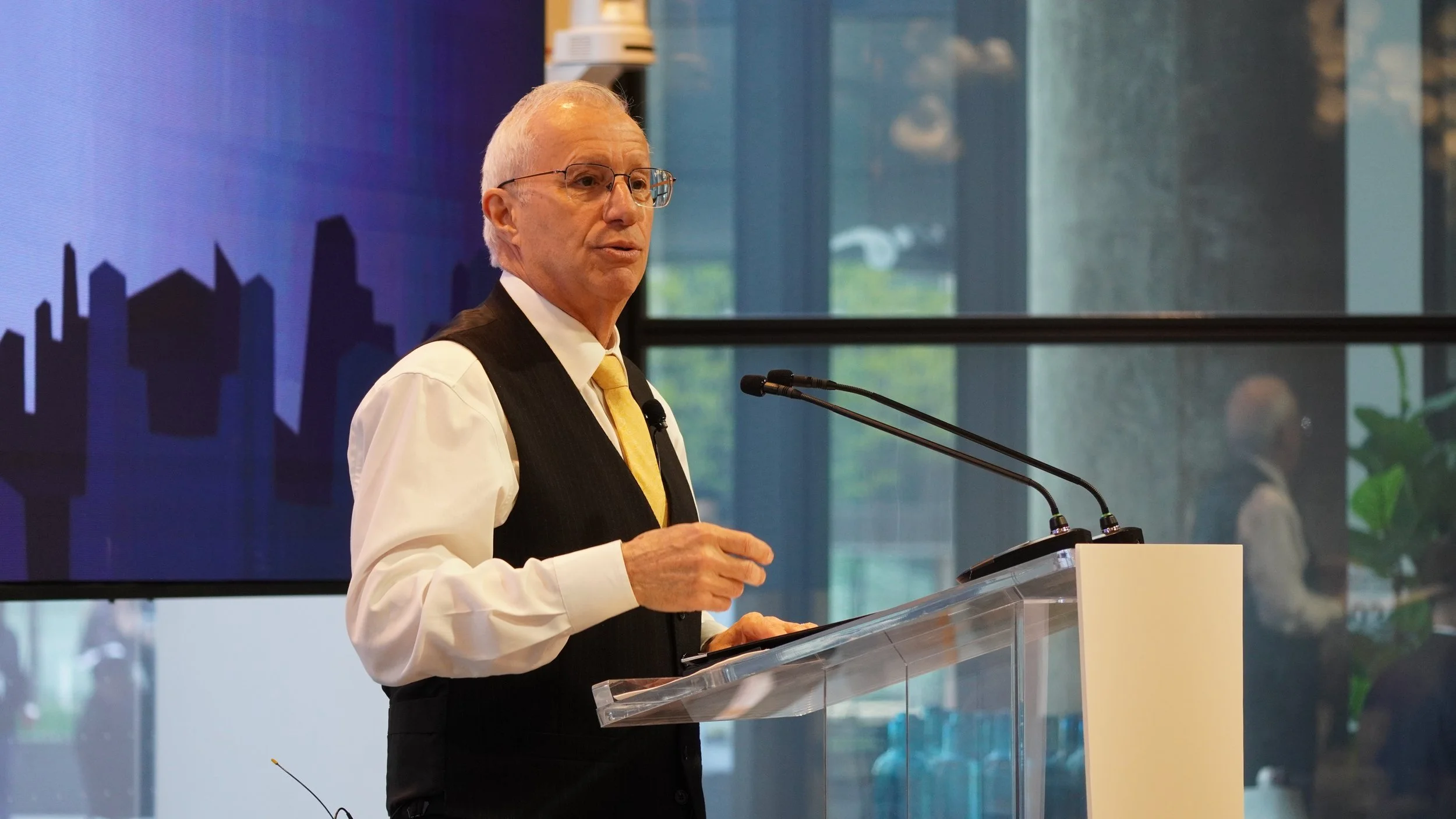 An older man with glasses and gray hair, dressed in a white shirt, black vest, and yellow tie, standing at a podium giving a speech.