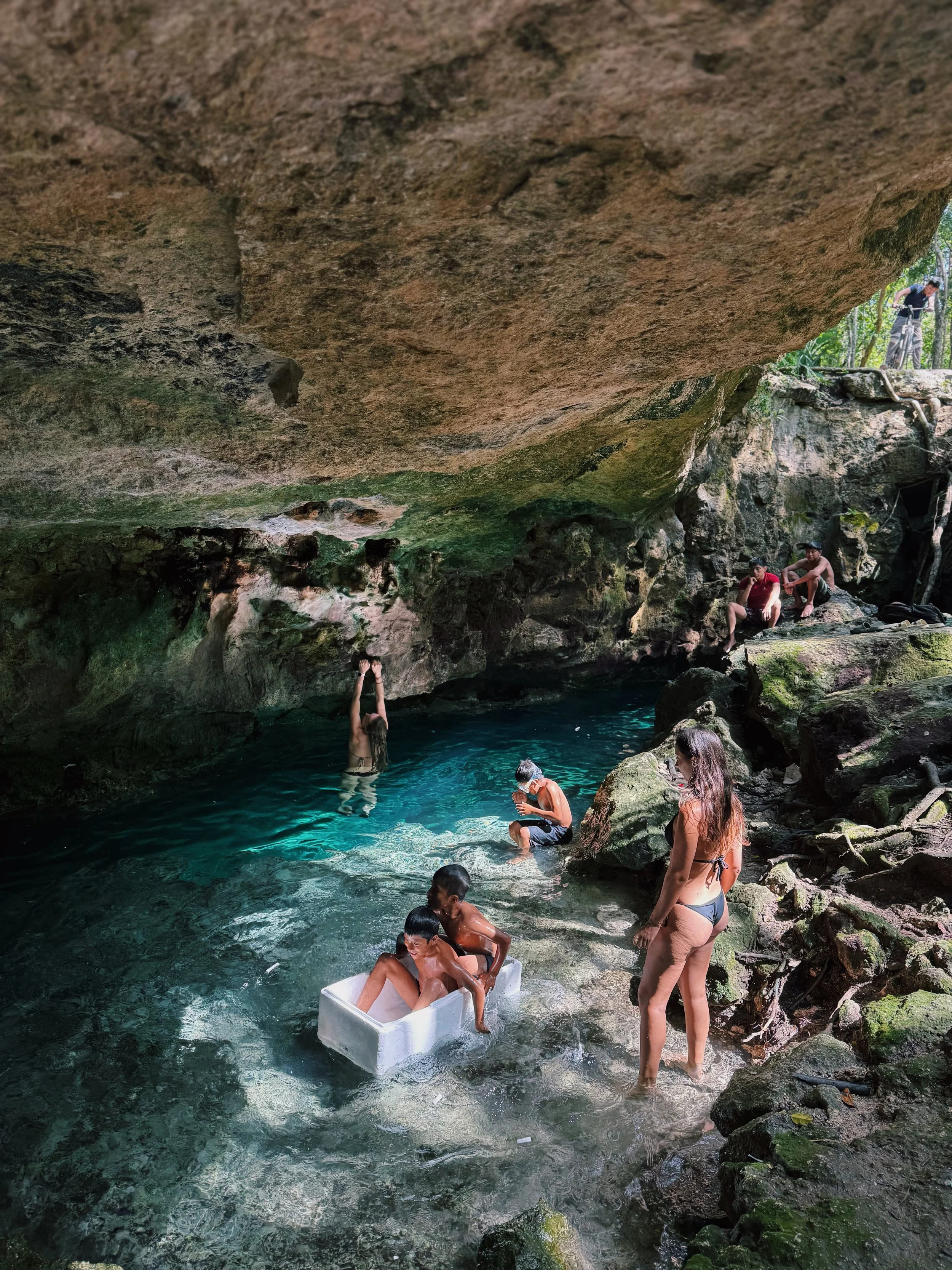 People swimming and relaxing in a natural rocky pool under a large overhanging rock, surrounded by mossy stones and trees.