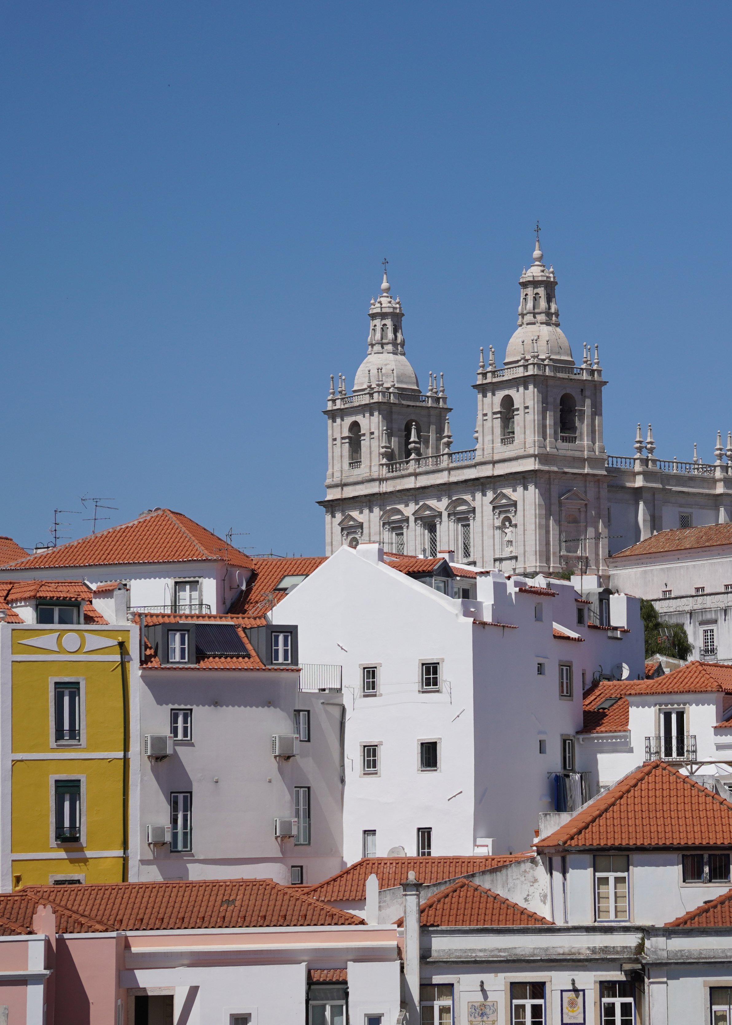 View of white and colored buildings with red tiled roofs under a clear blue sky, featuring a historic church with twin towers in the background.