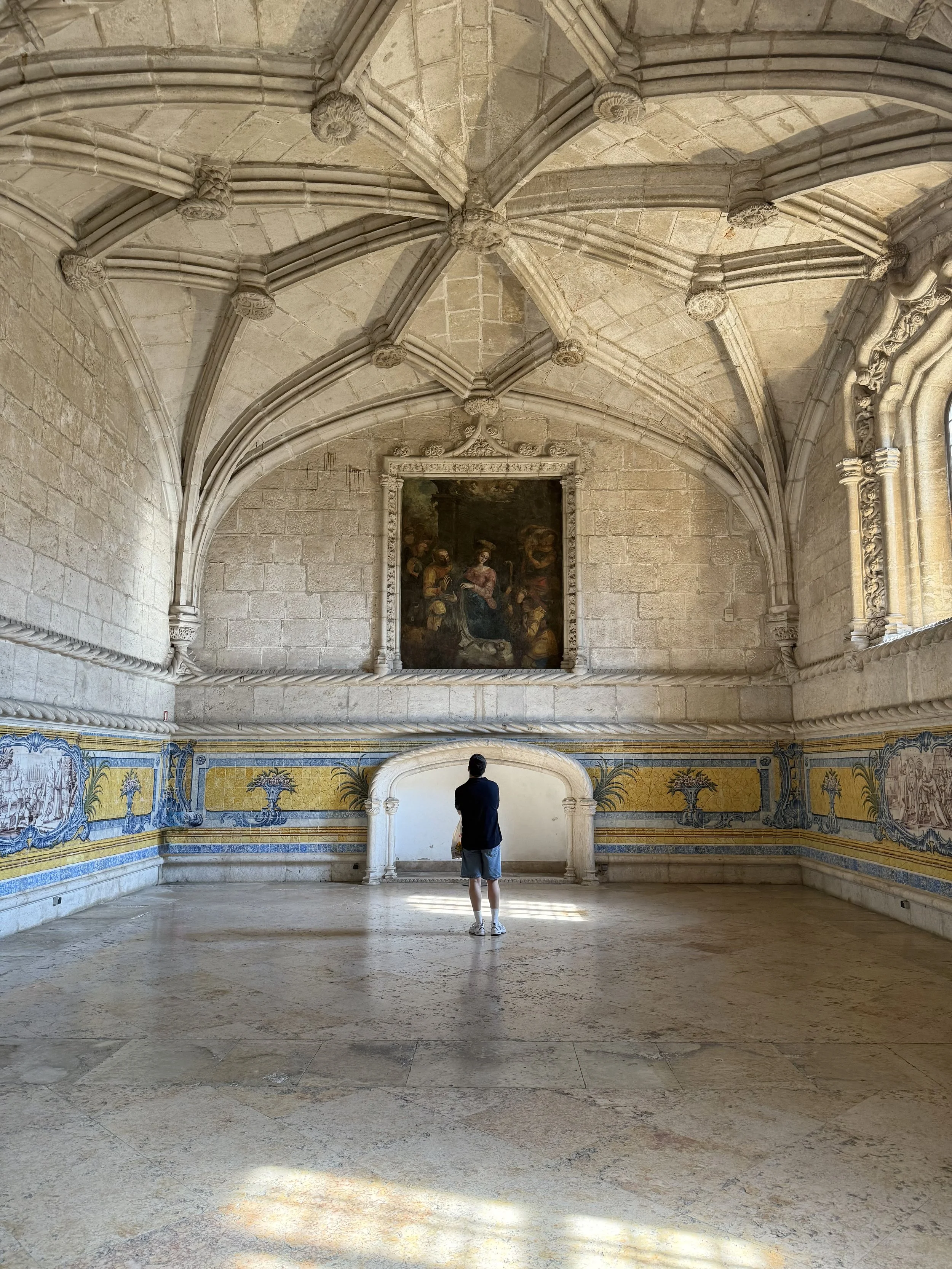 A person stands in a large, ornate hall with high, vaulted stone ceilings and decorative architectural details. They are facing a painting on the wall, which is framed and elevated. The lower part of the walls has colorful mosaic or tile artwork, and