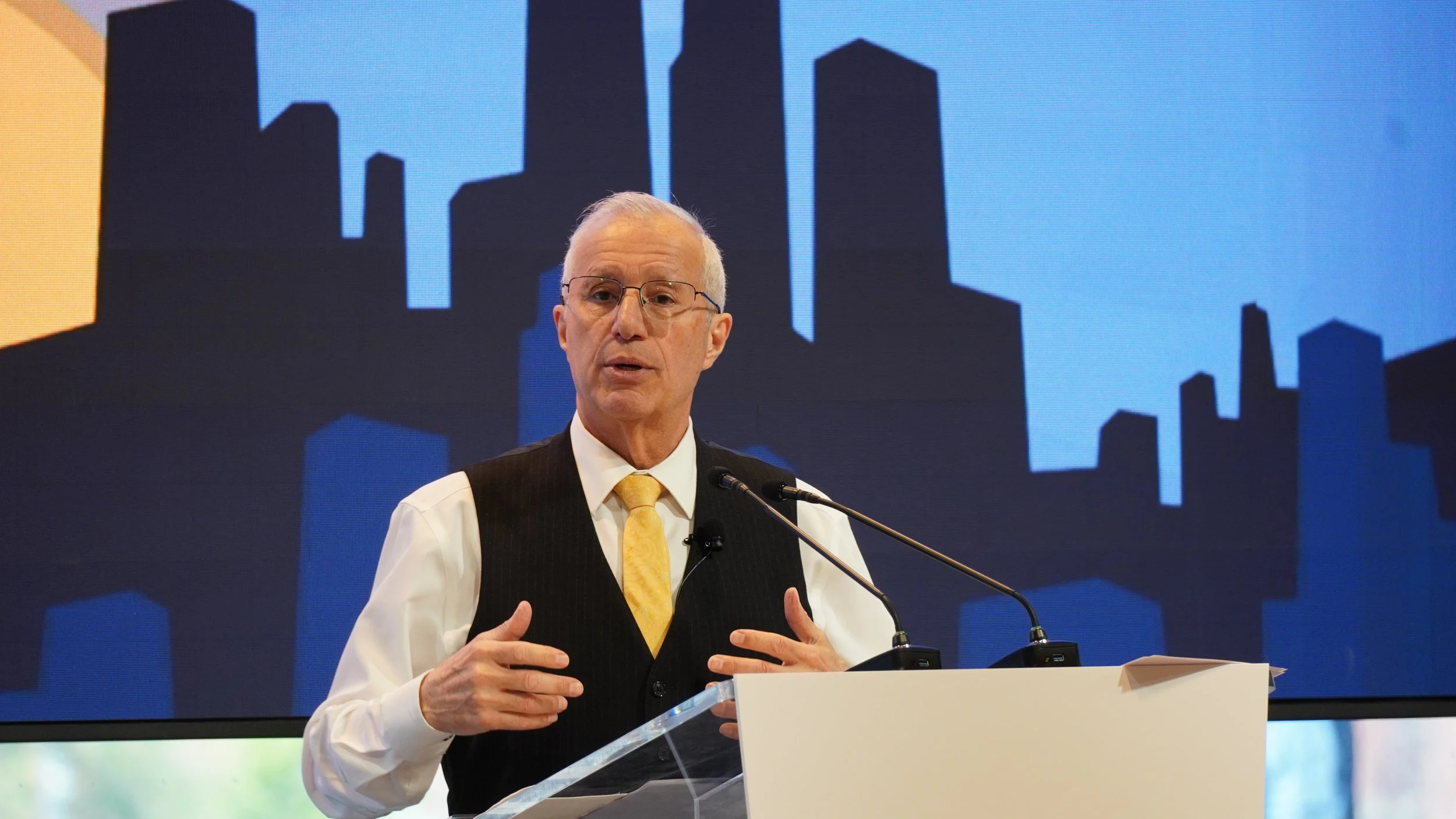 A man with glasses, white hair, and light skin standing at a podium with a microphone, wearing a white shirt, yellow tie, and dark vest, speaking in front of a large digital screen showing a blue urban city skyline silhouette.