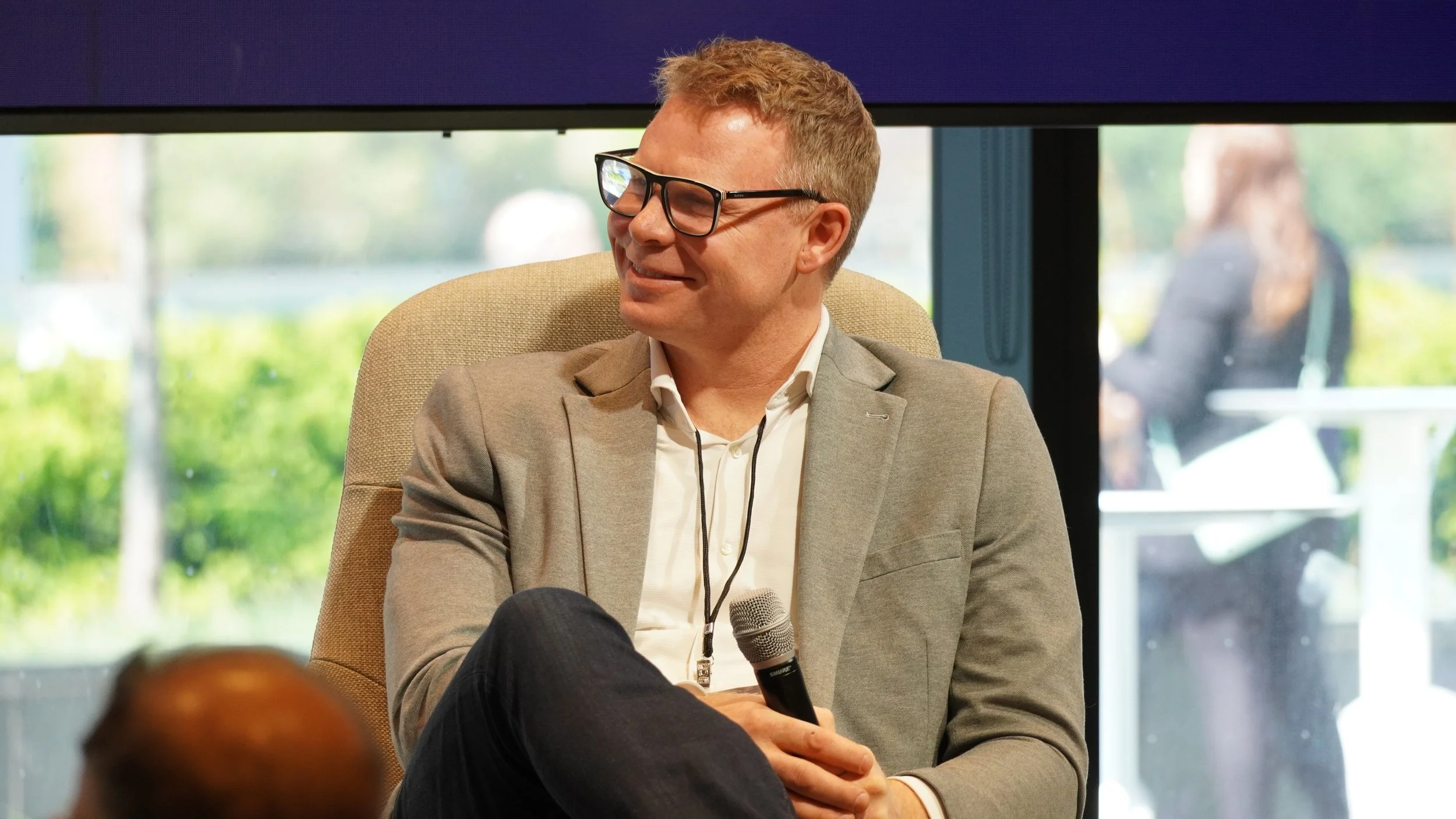 A man with glasses, wearing a gray blazer and white shirt, sitting on a beige chair, smiling and holding a microphone during a discussion or presentation, with a large window and a woman in the background.
