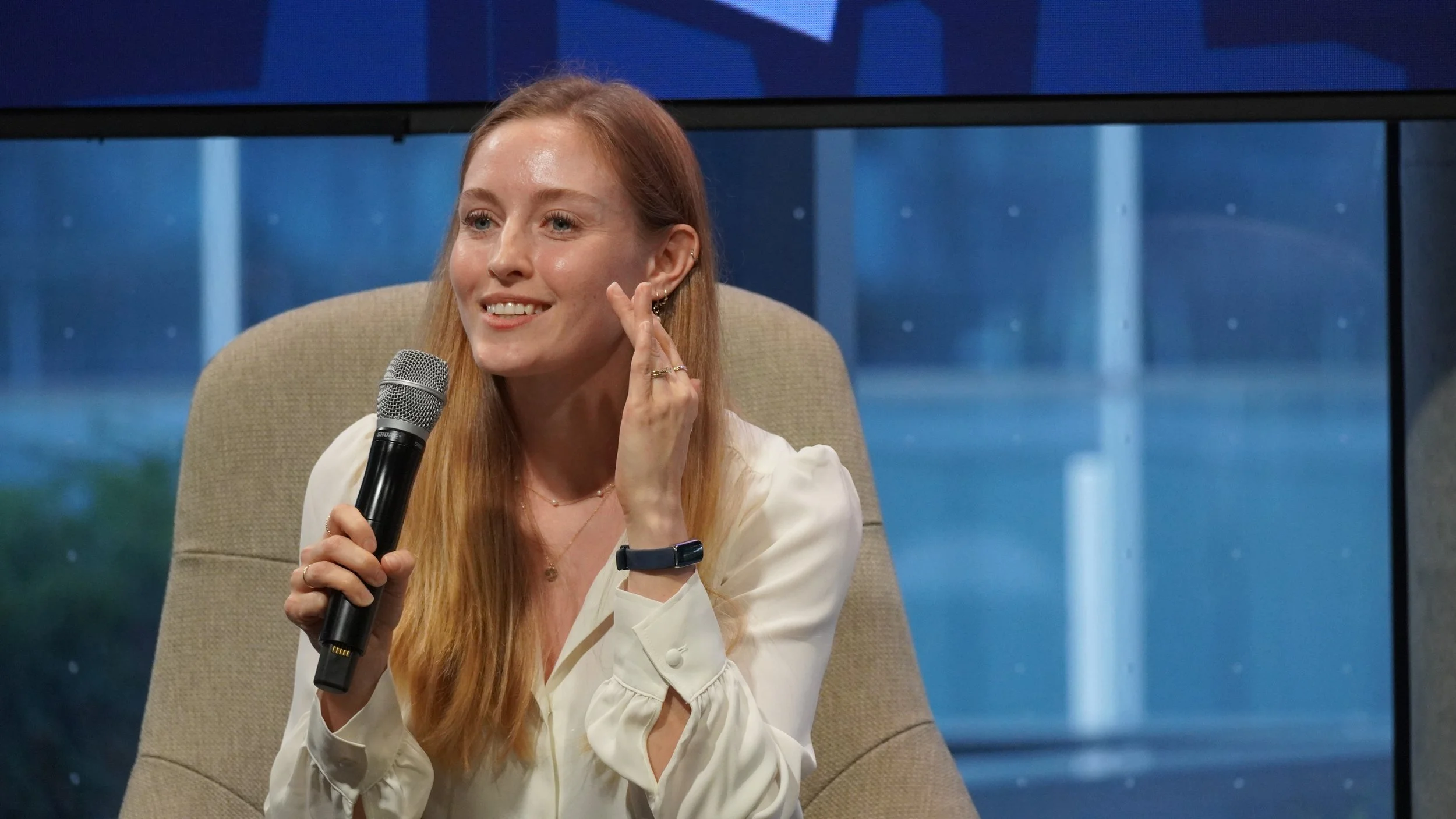 A woman with long red hair sitting on a beige chair, holding a microphone, smiling, wearing a white blouse, and touching her left ear, with a window and buildings in the background.