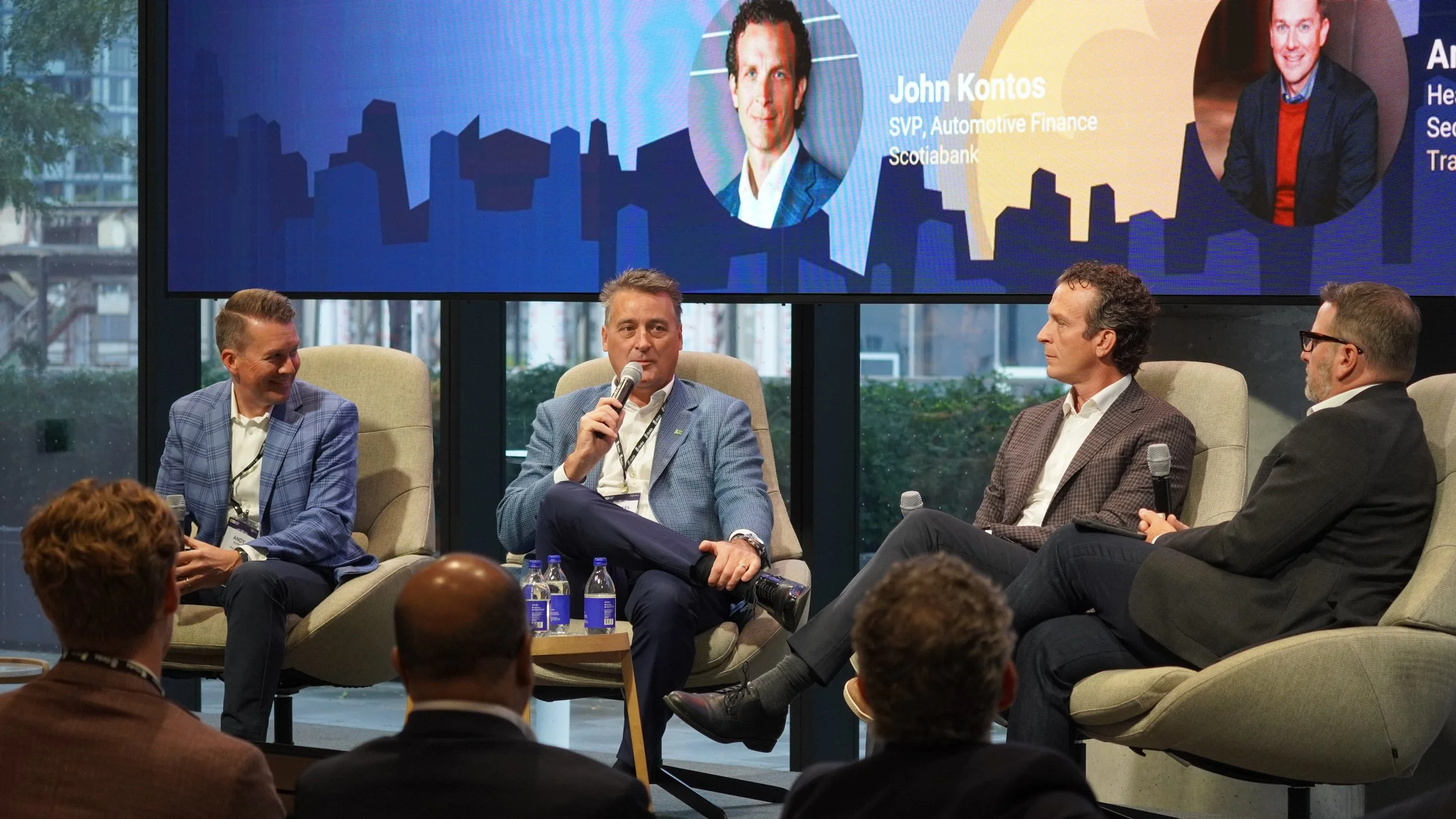 Panel discussion with four men seated in chairs, three holding microphones, in a modern conference room. A large screen behind them displays headshots and names of panelists, including John Kontos.
