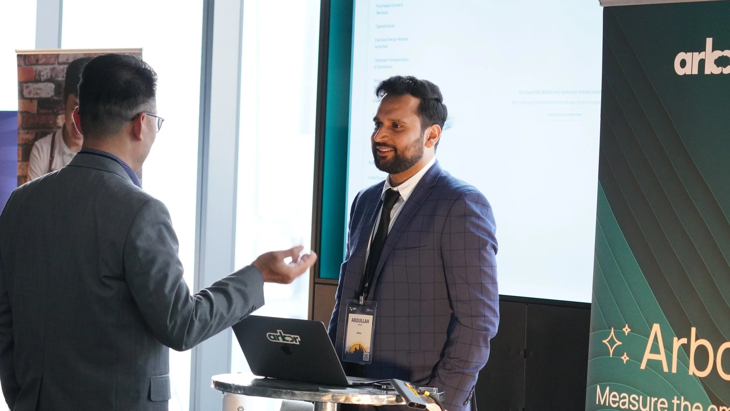 Two men in business suits engaging in a conversation at a professional event. One man is facing away from the camera with gray hair and glasses, while the other man, who is smiling, has dark hair and a beard. They're standing next to a table with a l