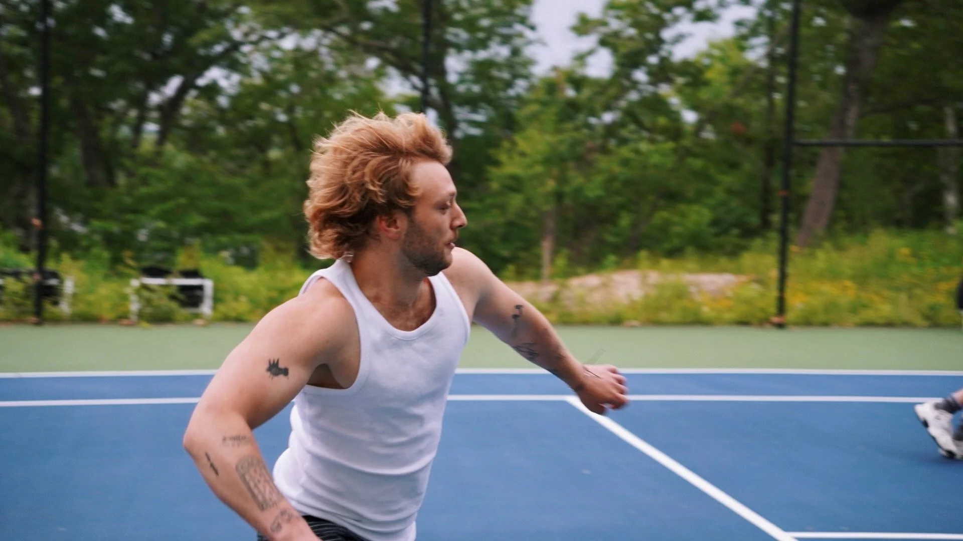 A man with long red hair and tattoos on his arms running on a blue outdoor tennis court surrounded by green trees.