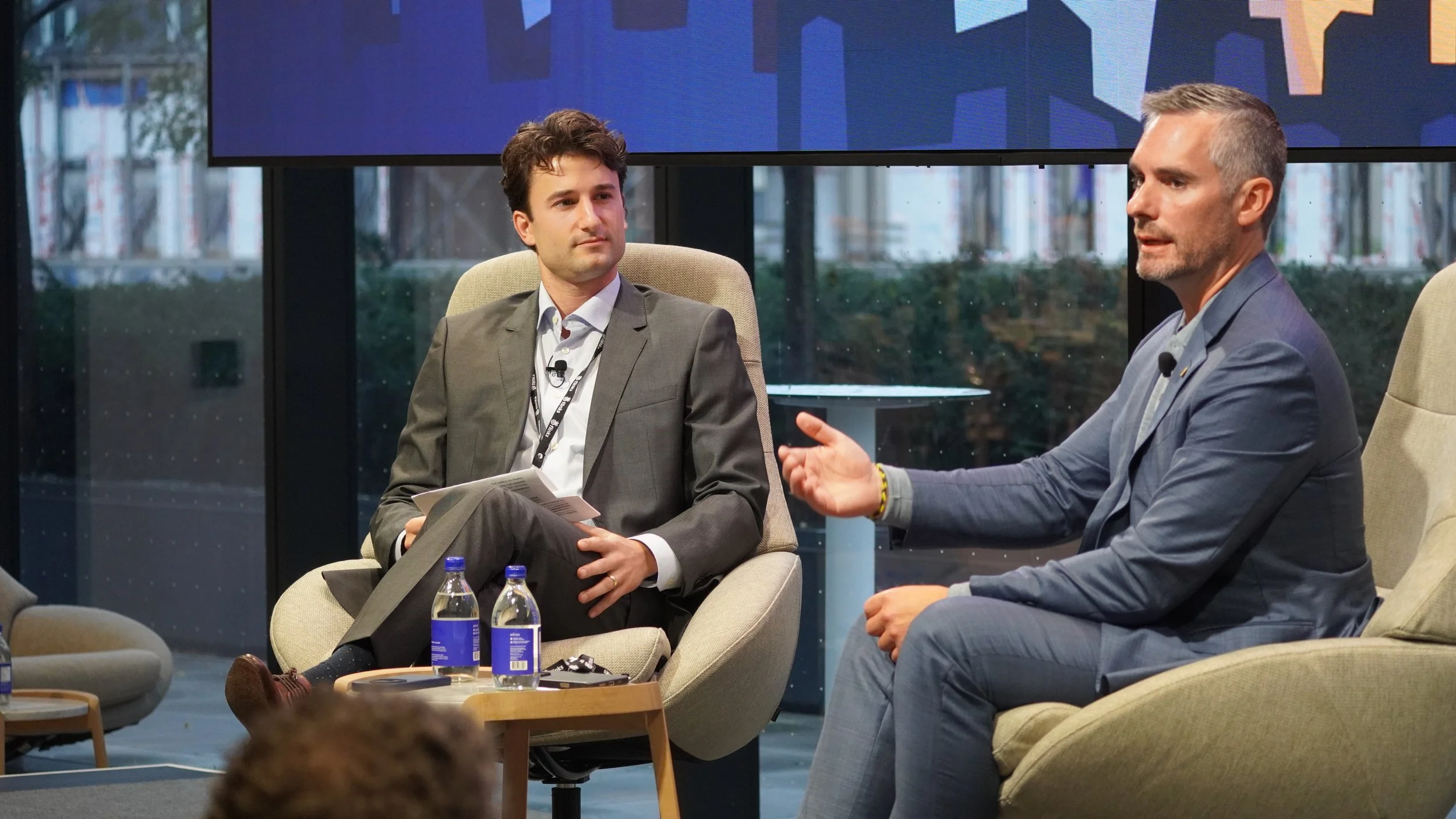 Two men in business suits engaged in a discussion on stage during a conference or panel. One man is speaking, the other is listening, both seated in cushioned chairs. There are bottled waters on a small table between them and a large digital screen i