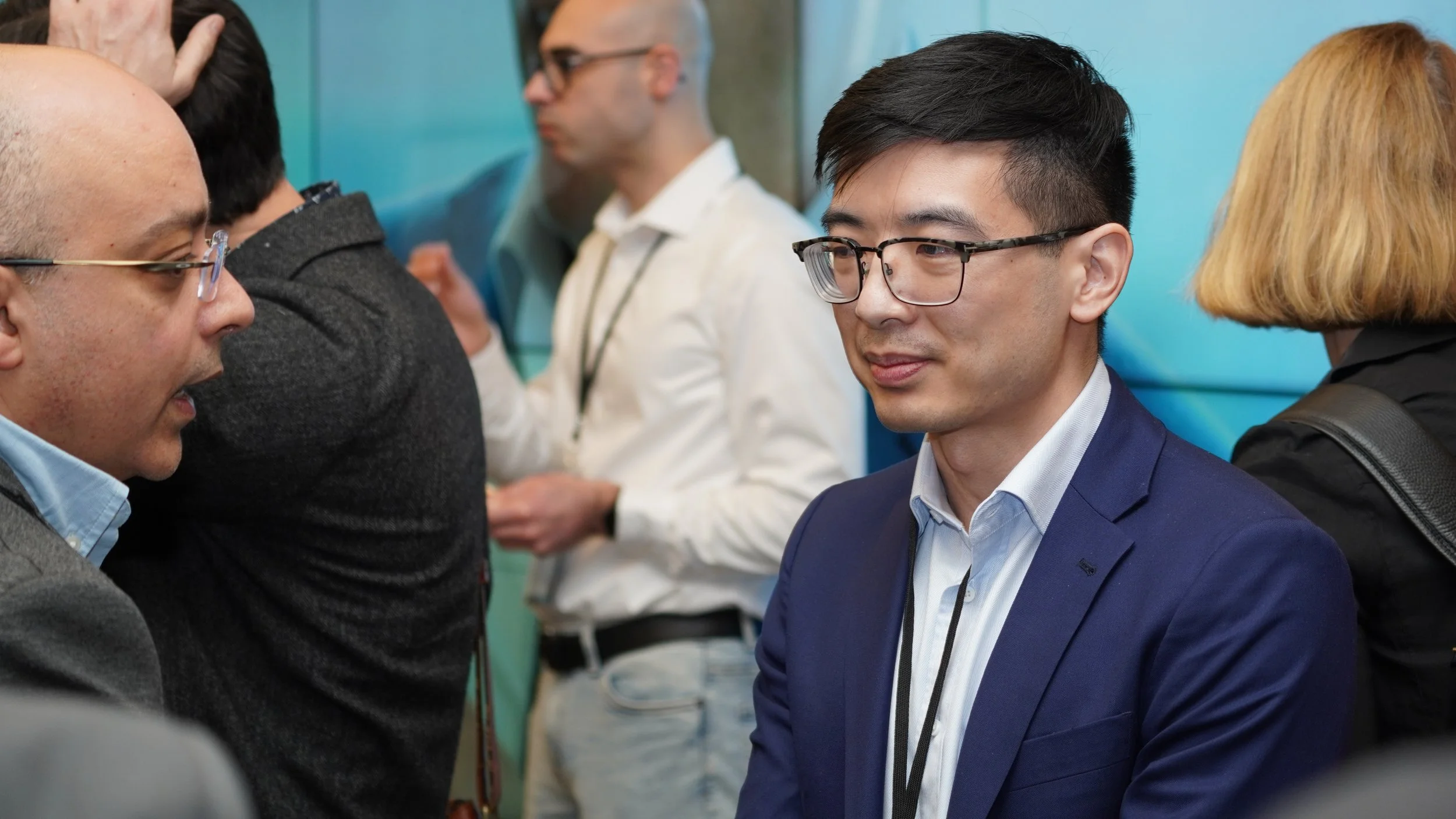A man in a blue suit with glasses and black hair is talking with a person not visible in the photo. In the background, three other people are engaged in conversations at a professional event.
