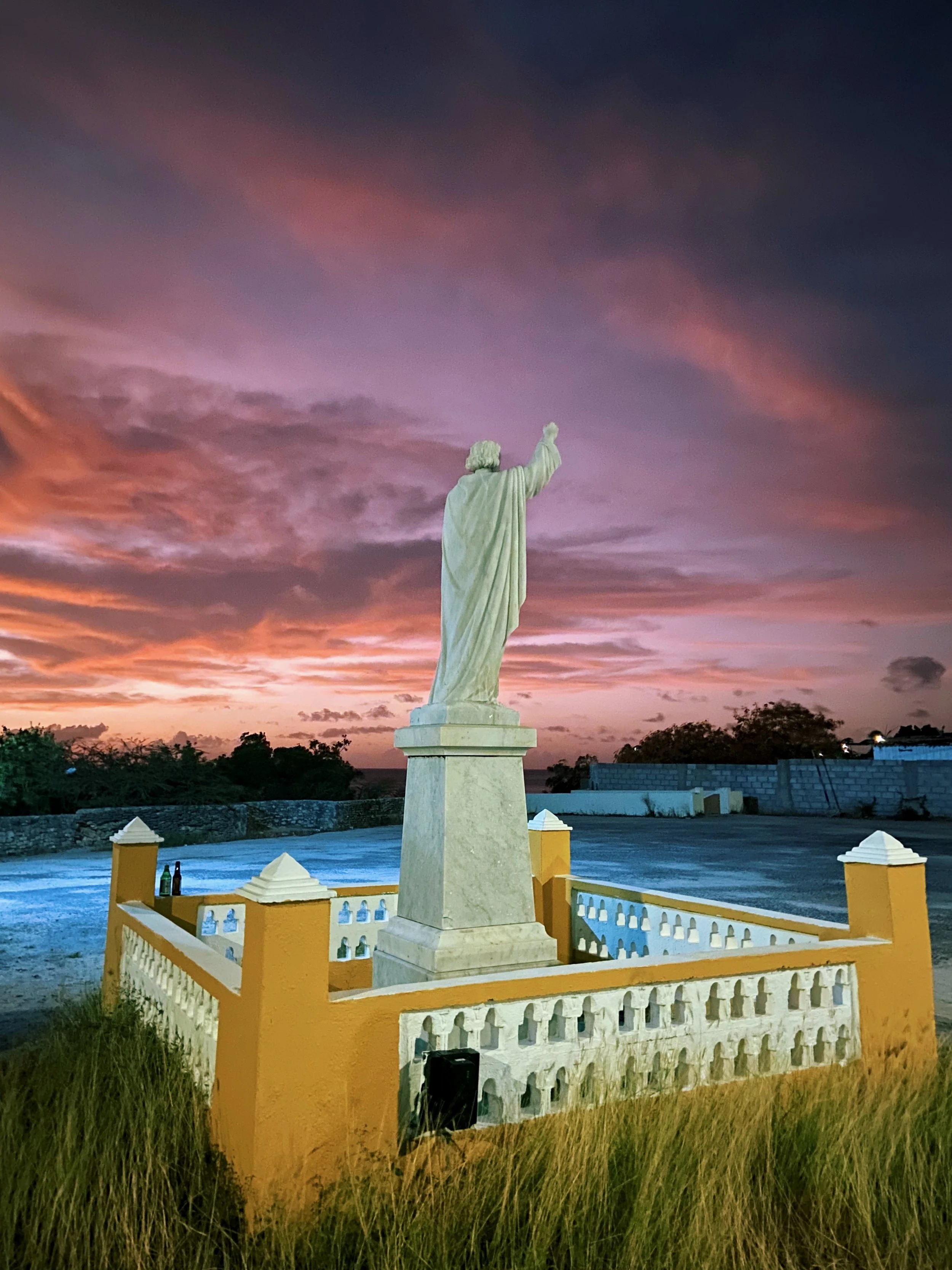 Statue of a person with one arm raised, situated on a yellow and white platform, with a colorful sunset sky in the background.