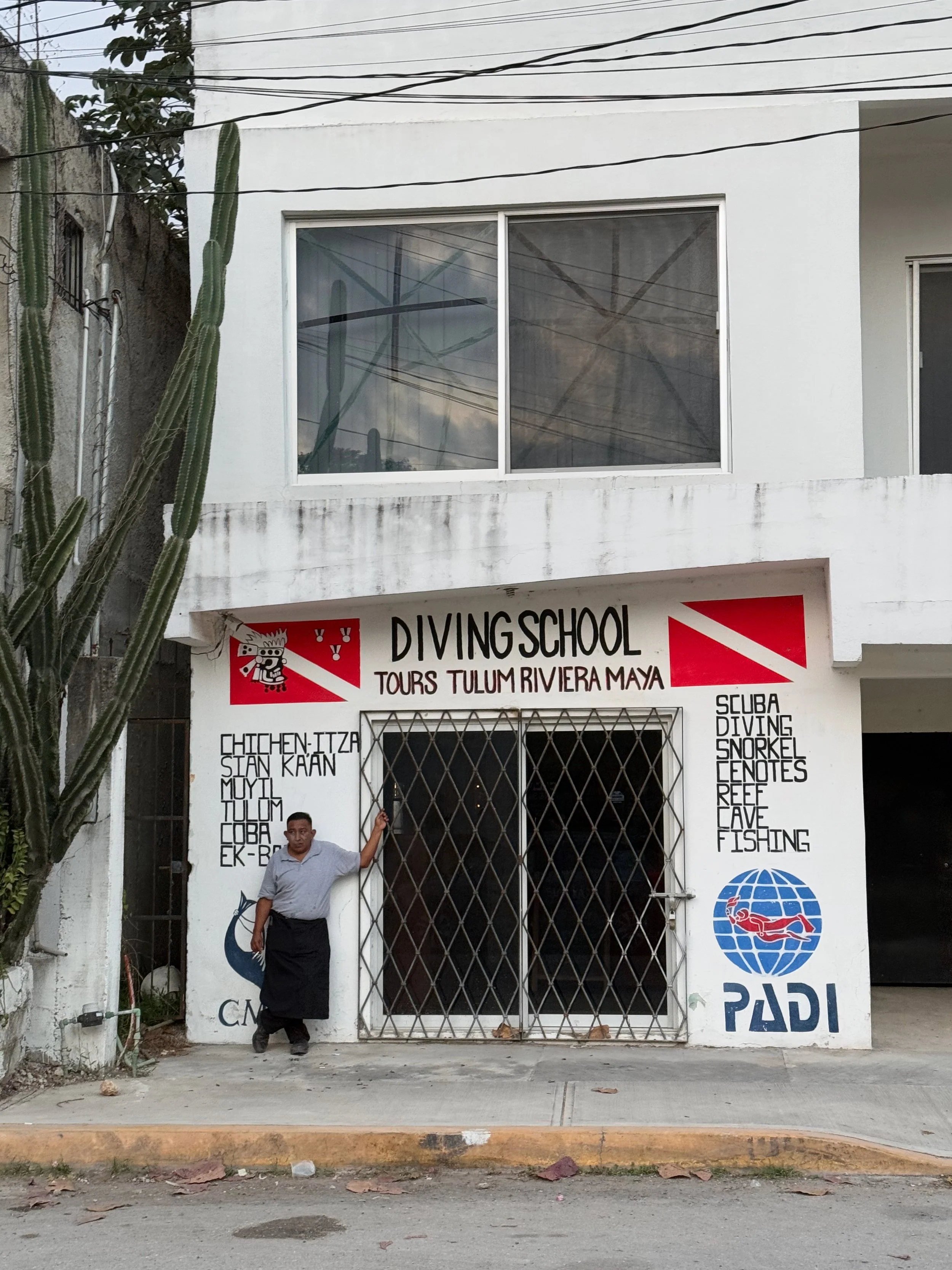 Front view of a white building with a sign reading 'Divingschool Tours Tulum Riviera Maya' and logos for scuba diving, snorkeling, cenotes, reef, cave, and fishing. A man in a gray shirt and black apron stands in front of the gated entrance.