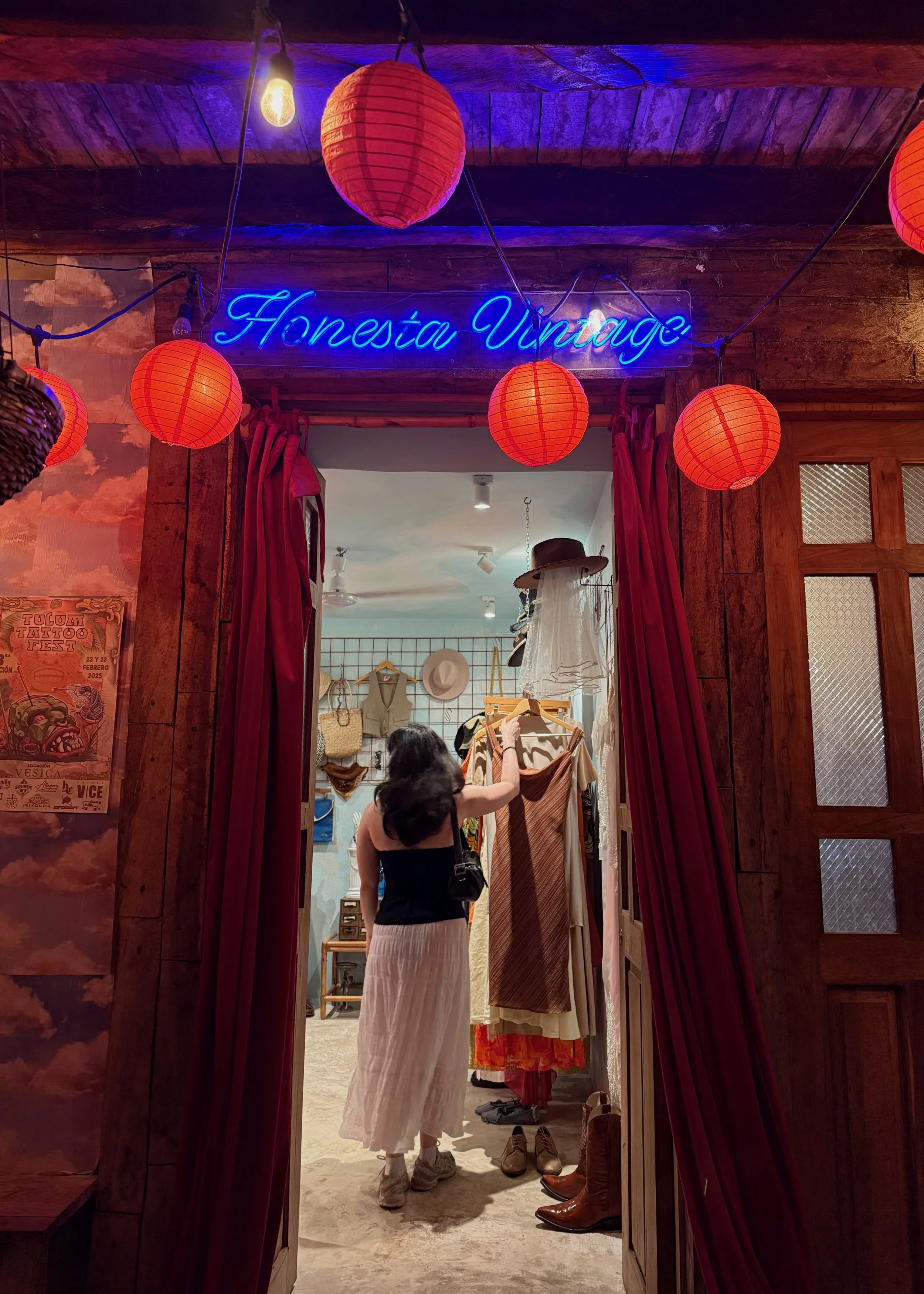 A person shopping for clothing inside a store with a sign that reads 'Honest Vintage'; red lanterns and warm lighting decor.