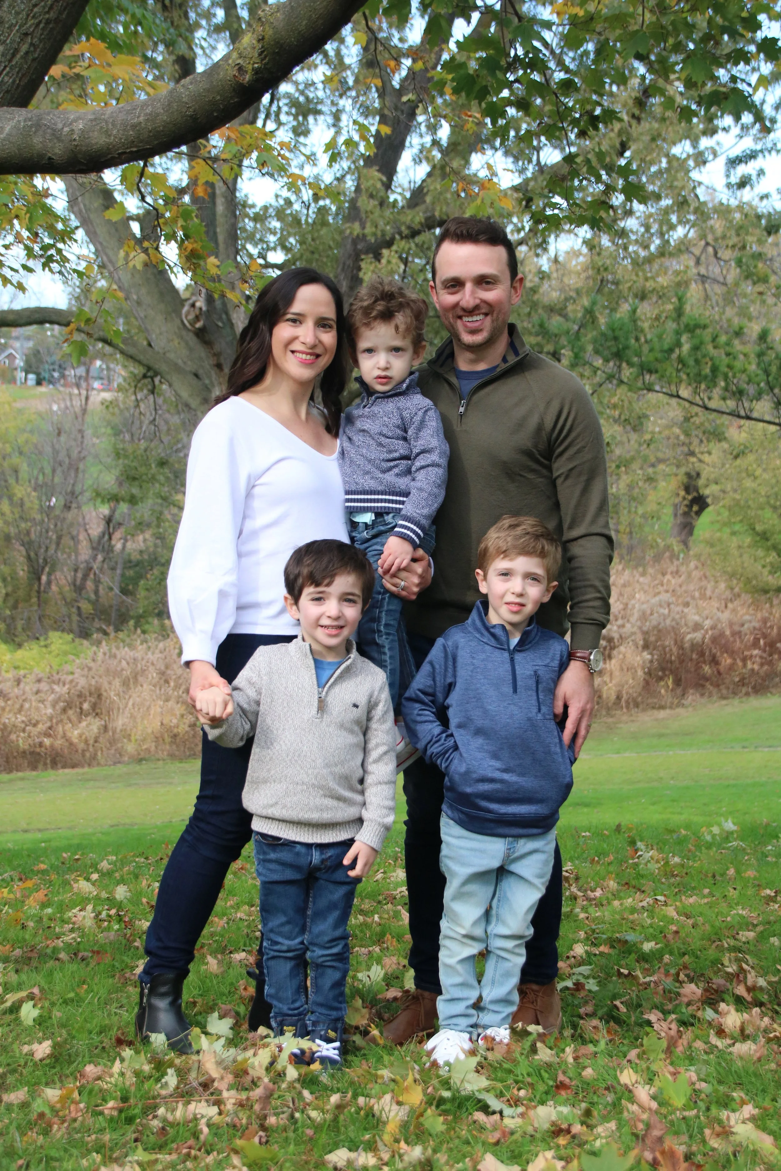 A family of six standing outdoors in a park with green grass, trees, and autumn leaves.