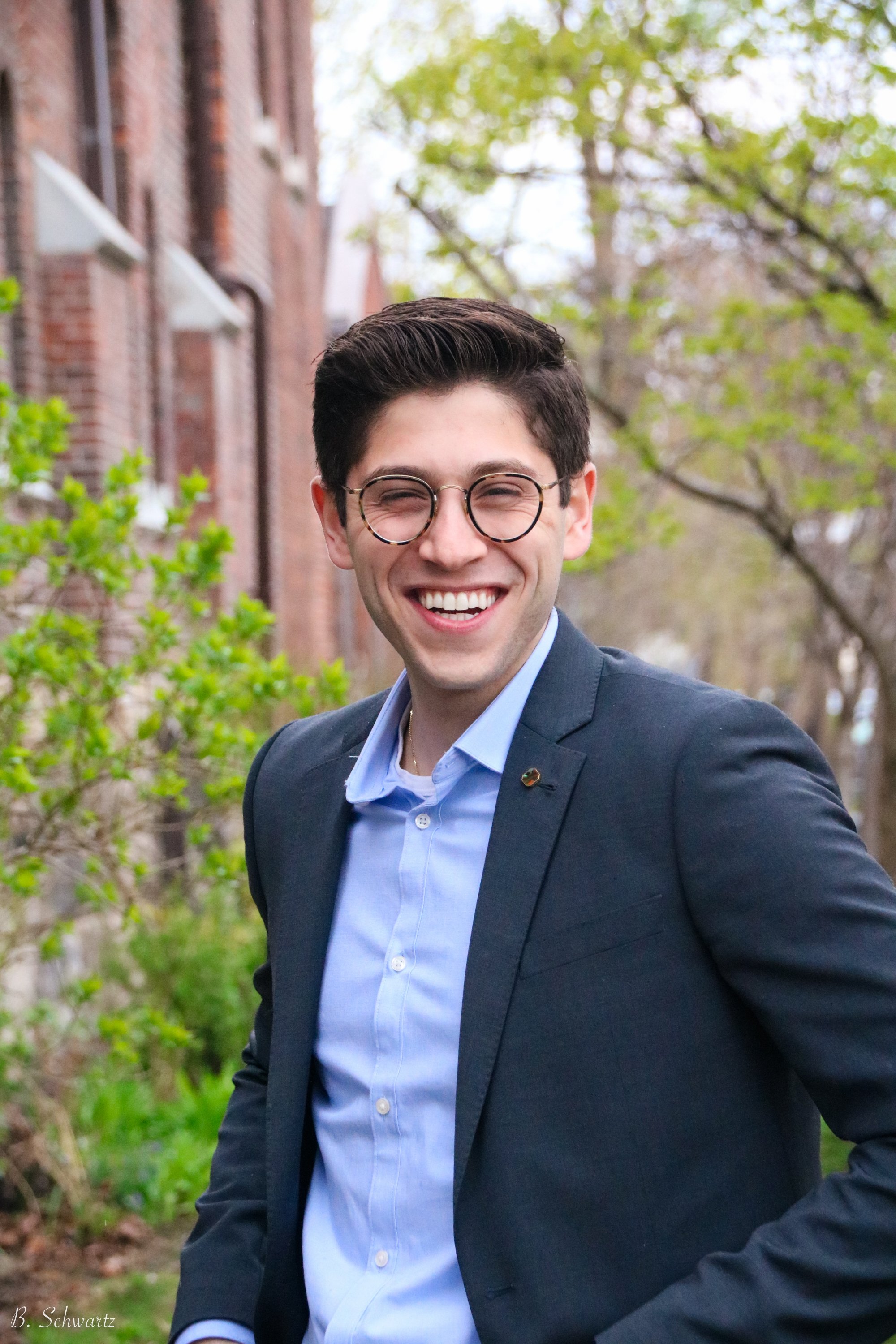 A young man in a suit and glasses smiling outdoors near trees and a brick building.