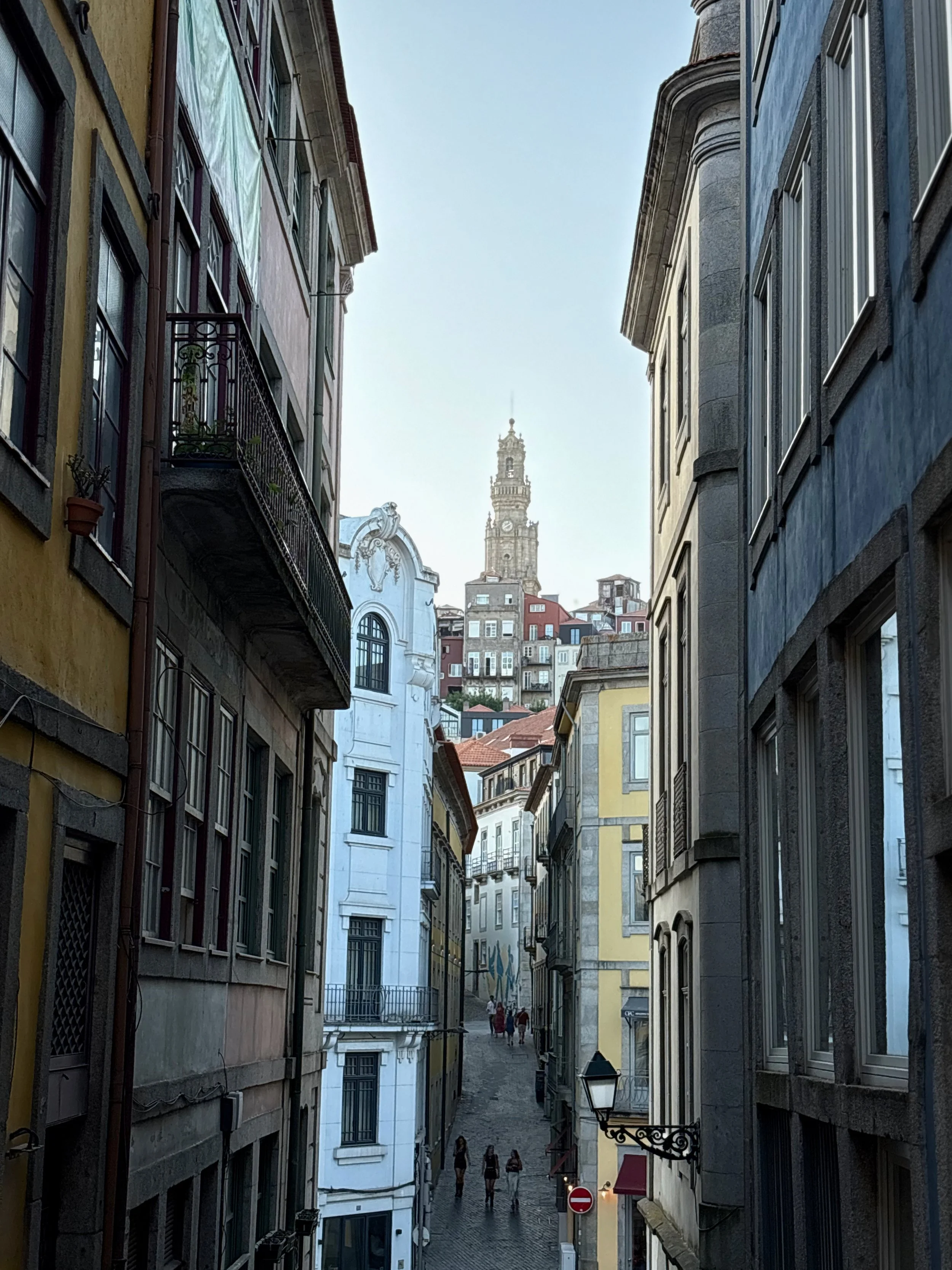 A narrow cobblestone street in a European city lined with colorful old buildings, with a view of a tall tower in the background.