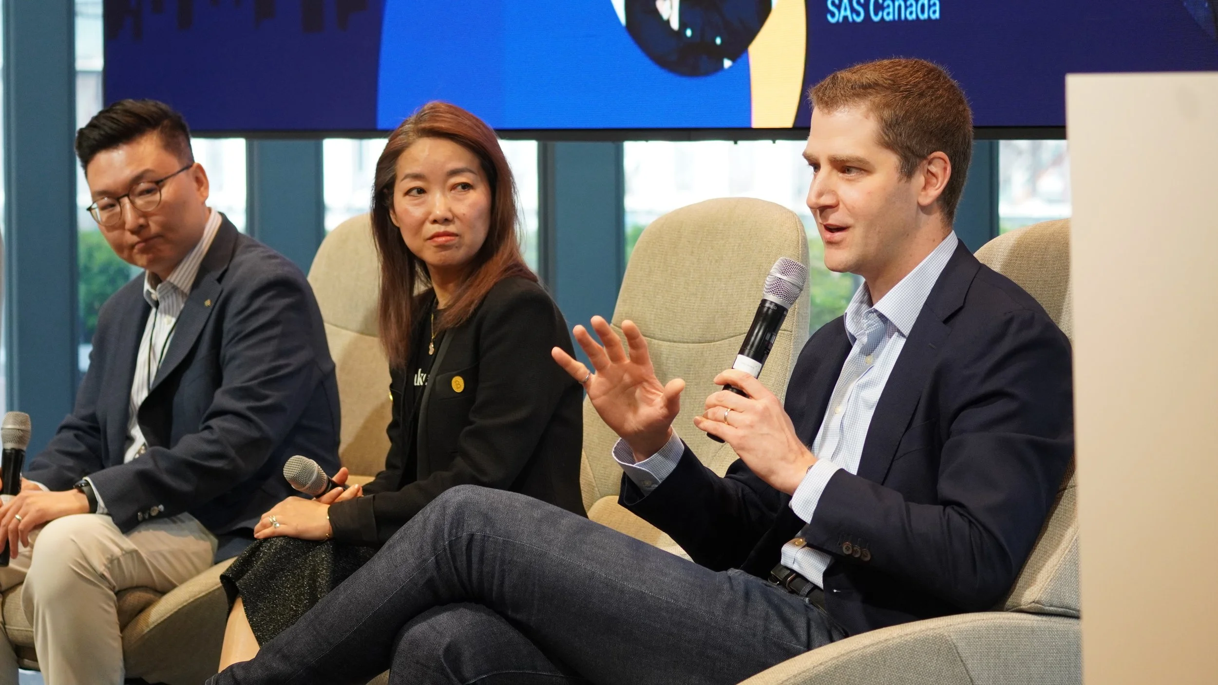 A panel of three people sitting in beige chairs, participating in a discussion at an event. The man on the right is speaking into a microphone, gesturing with his hand. The woman in the middle and the man on the left are listening. The background sho