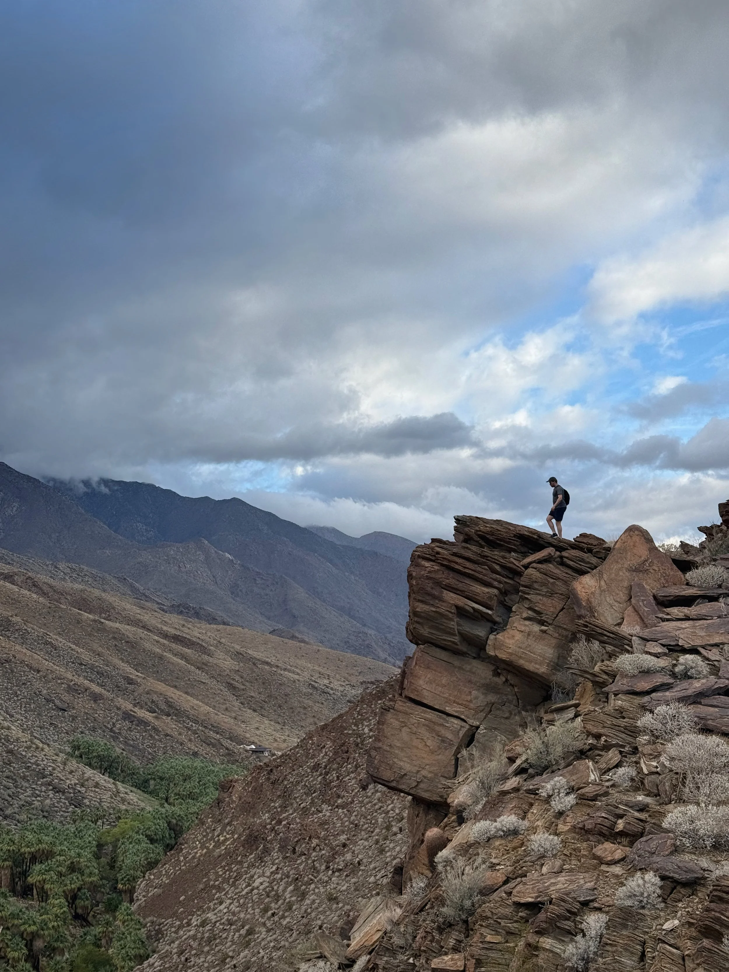 A person standing on the edge of a rocky cliff in a mountainous desert landscape with cloudy sky.
