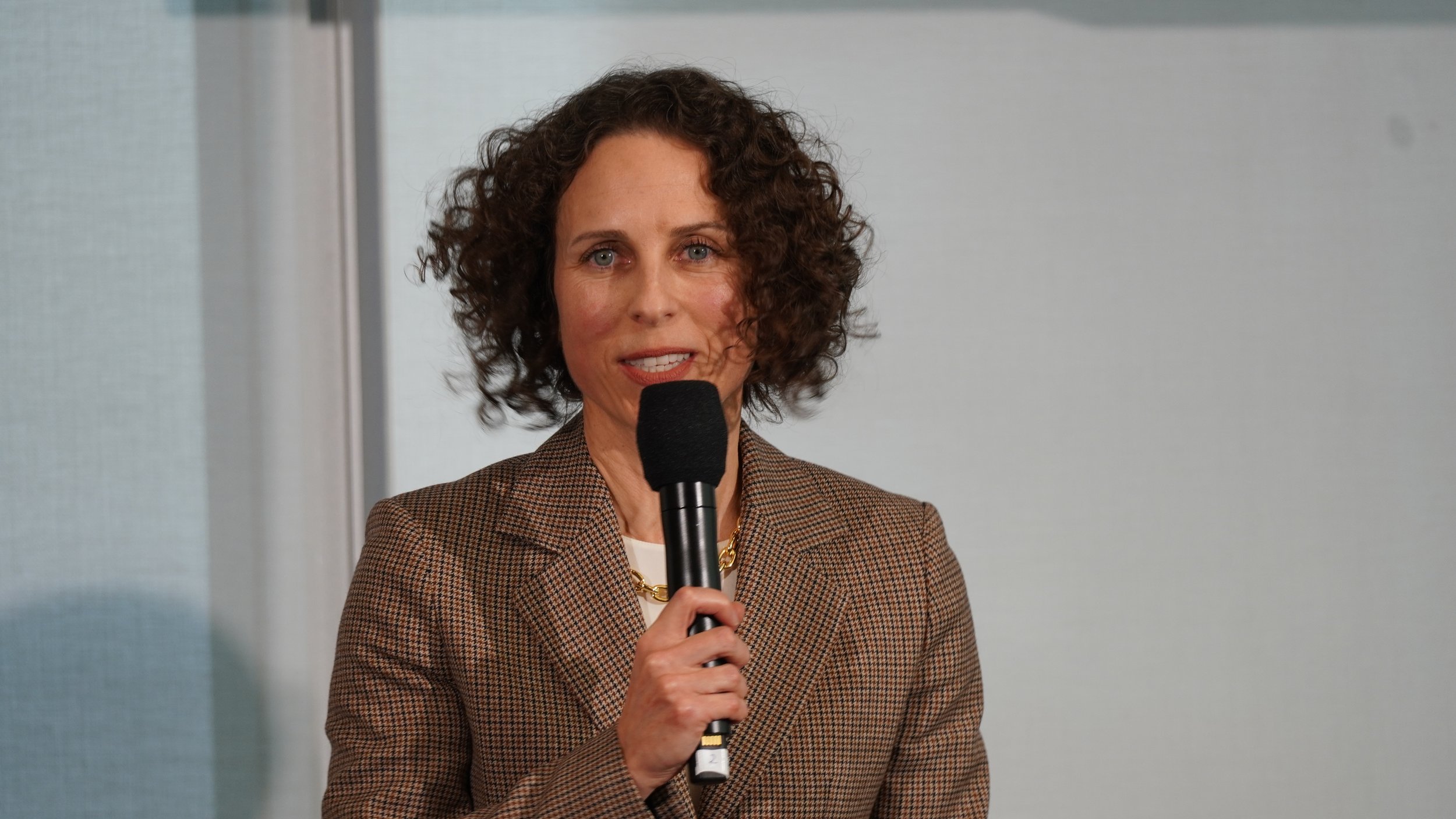 A woman with curly brown hair holding a microphone and speaking.
