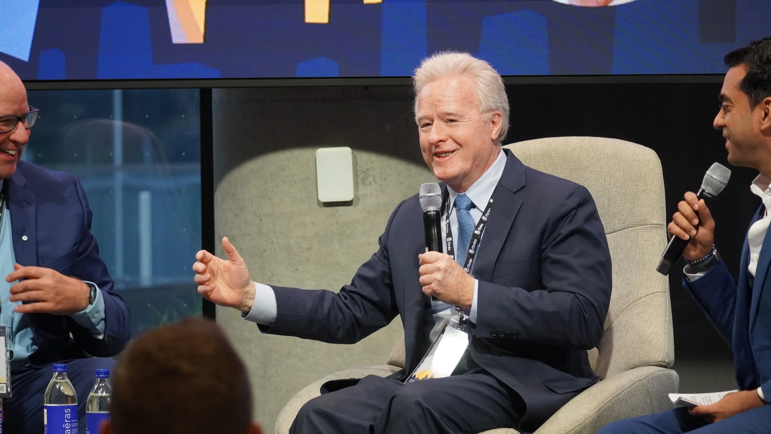 Three men in suits participating in a panel discussion, two of them holding microphones. The man in the center, with white hair, is speaking and gesturing with his hand. There are water bottles on the table and a large screen behind them.