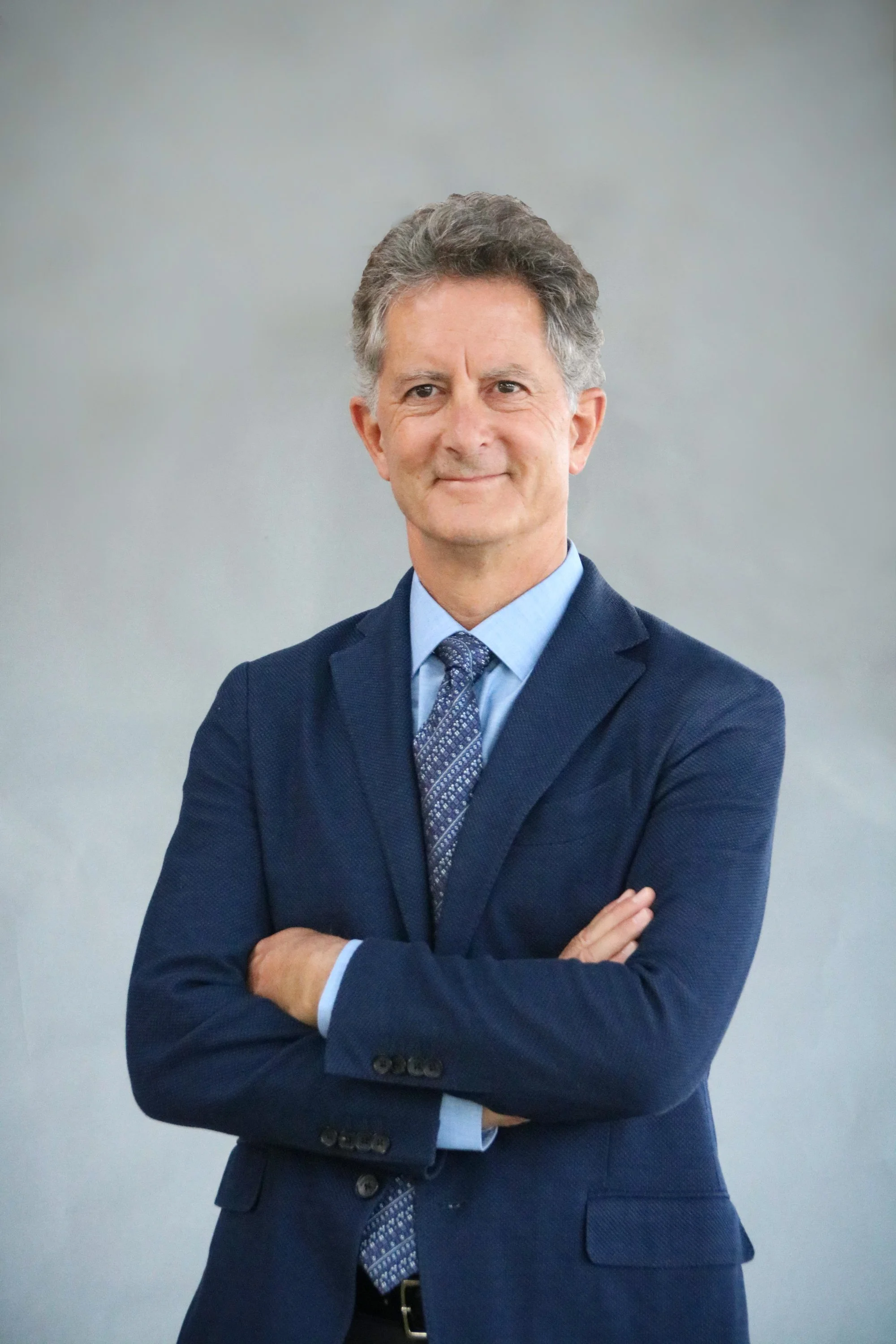 A professional middle-aged man in a navy blue suit, light blue shirt, and a patterned tie, standing with arms crossed and smiling softly against a neutral background.