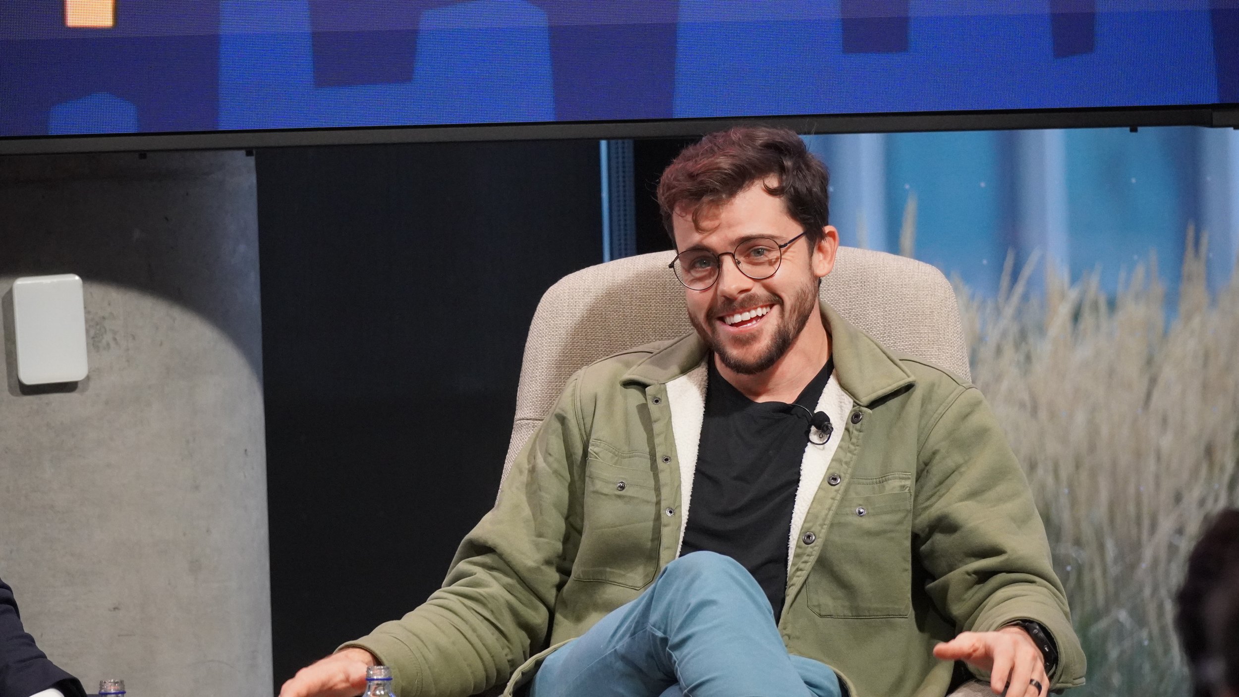 A smiling man with glasses and a beard, sitting in a beige chair, wearing a green jacket and black shirt, with a microphone clipped to his shirt. He is in an indoor setting with a large display screen above him and blurred plants in the background.