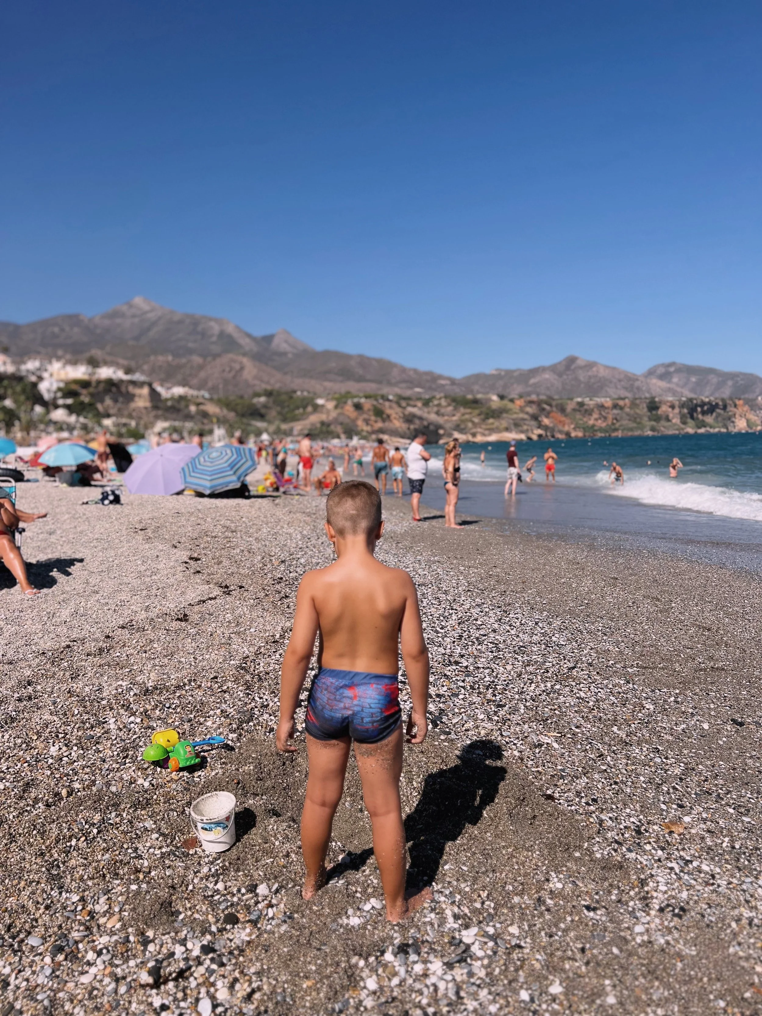 A young boy standing on a pebbly beach facing the ocean with people swimming and sunbathing, colorful umbrellas, mountains in the background, and a clear blue sky.
