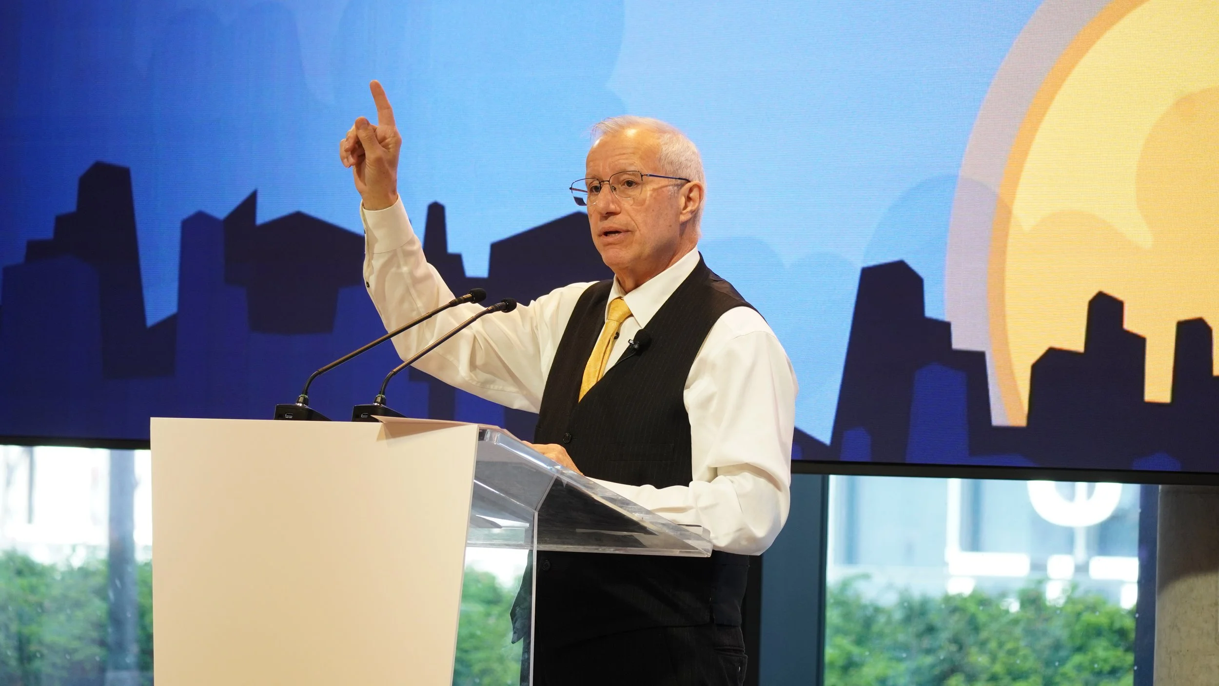 A man in a black vest and white shirt giving a speech at a podium with a colorful cityscape backdrop.