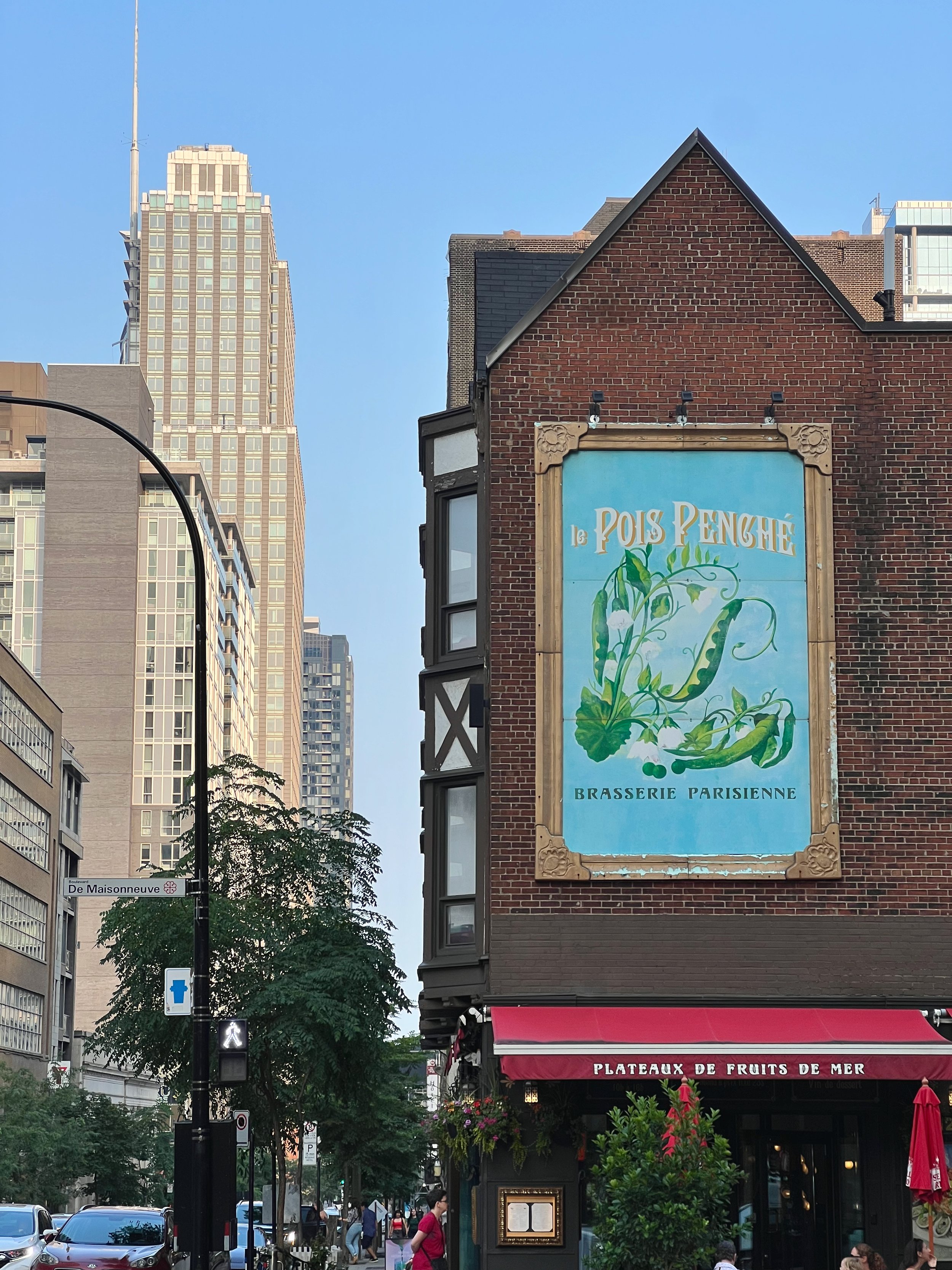 Street scene in an urban area with tall buildings, trees, a pedestrian crossing signal, and a restaurant with a red awning. There is a large sign on a brick building that reads 'Le Pois Penghé Brasserie Parisienne' with a painted artichoke, and a str