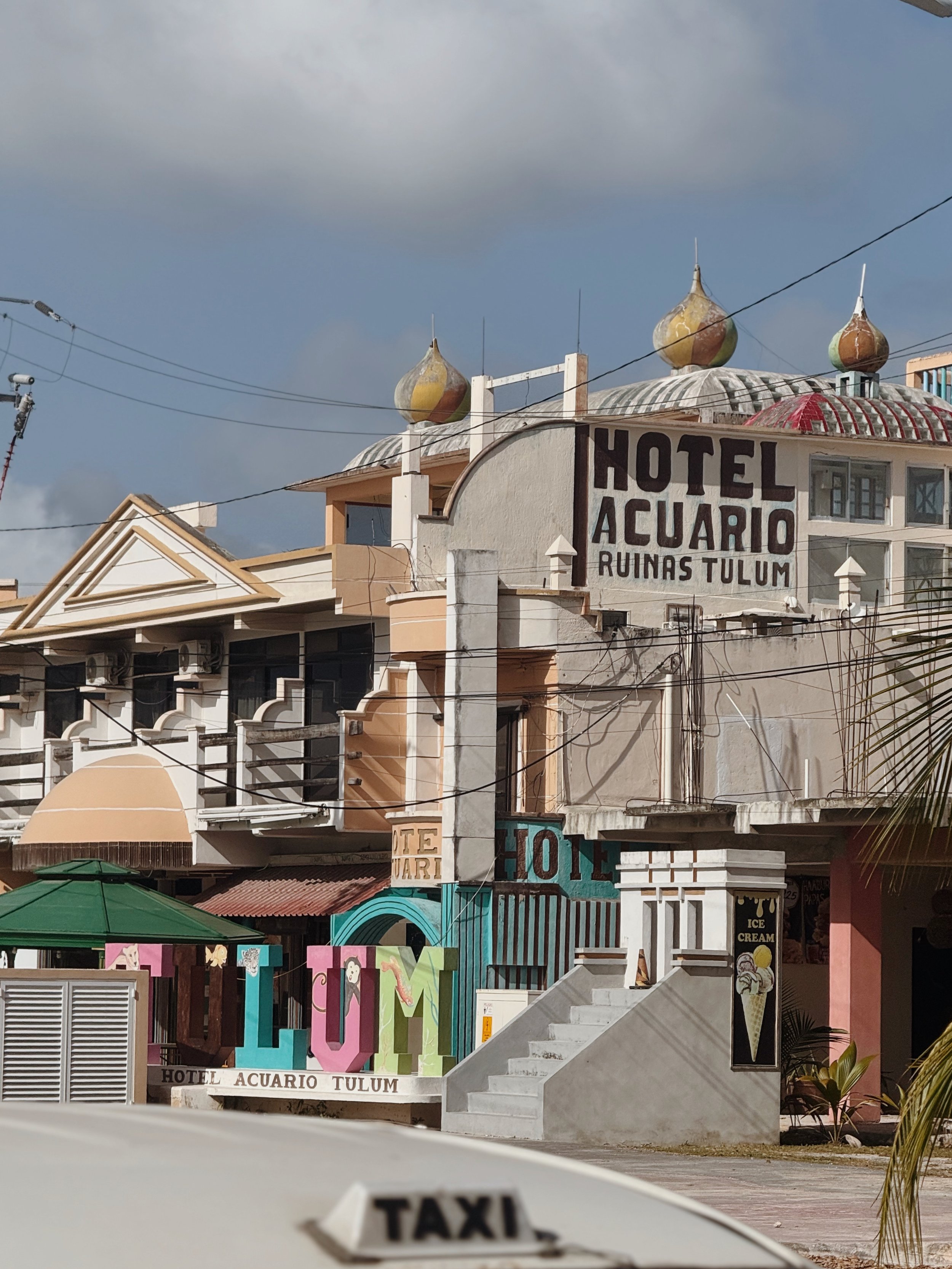 Buildings with colorful signage, including a hotel named Hotel Acuario Tulum, a large sign spelling LUM, and an ice cream sign, with a taxi in the foreground and a cloudy sky overhead.