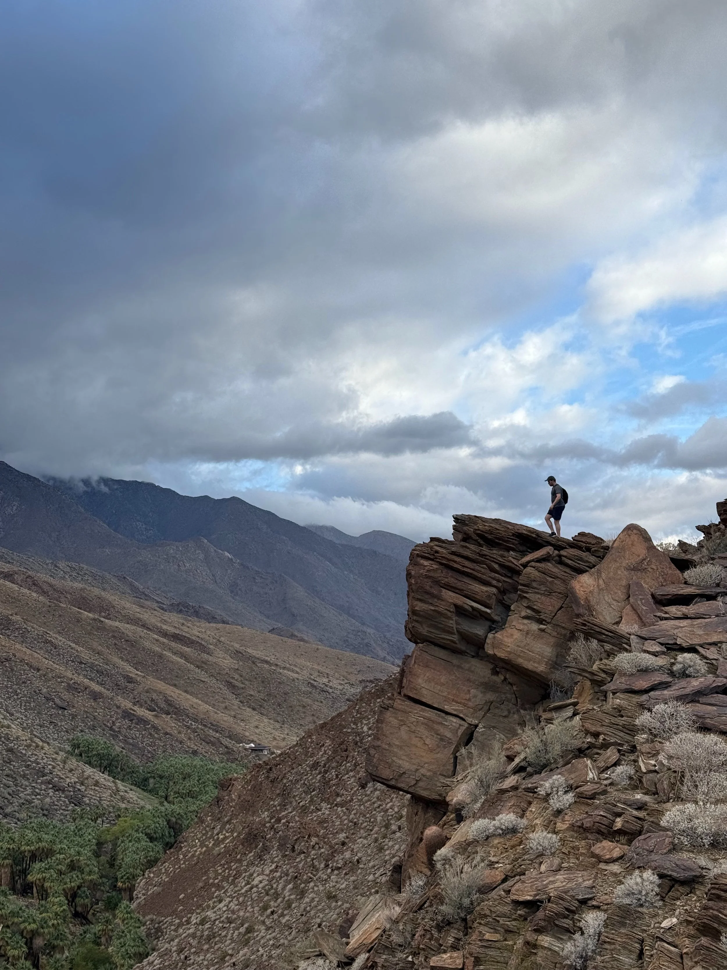 A person standing on the edge of a rocky cliff, overlooking a mountainous landscape with partly cloudy skies.